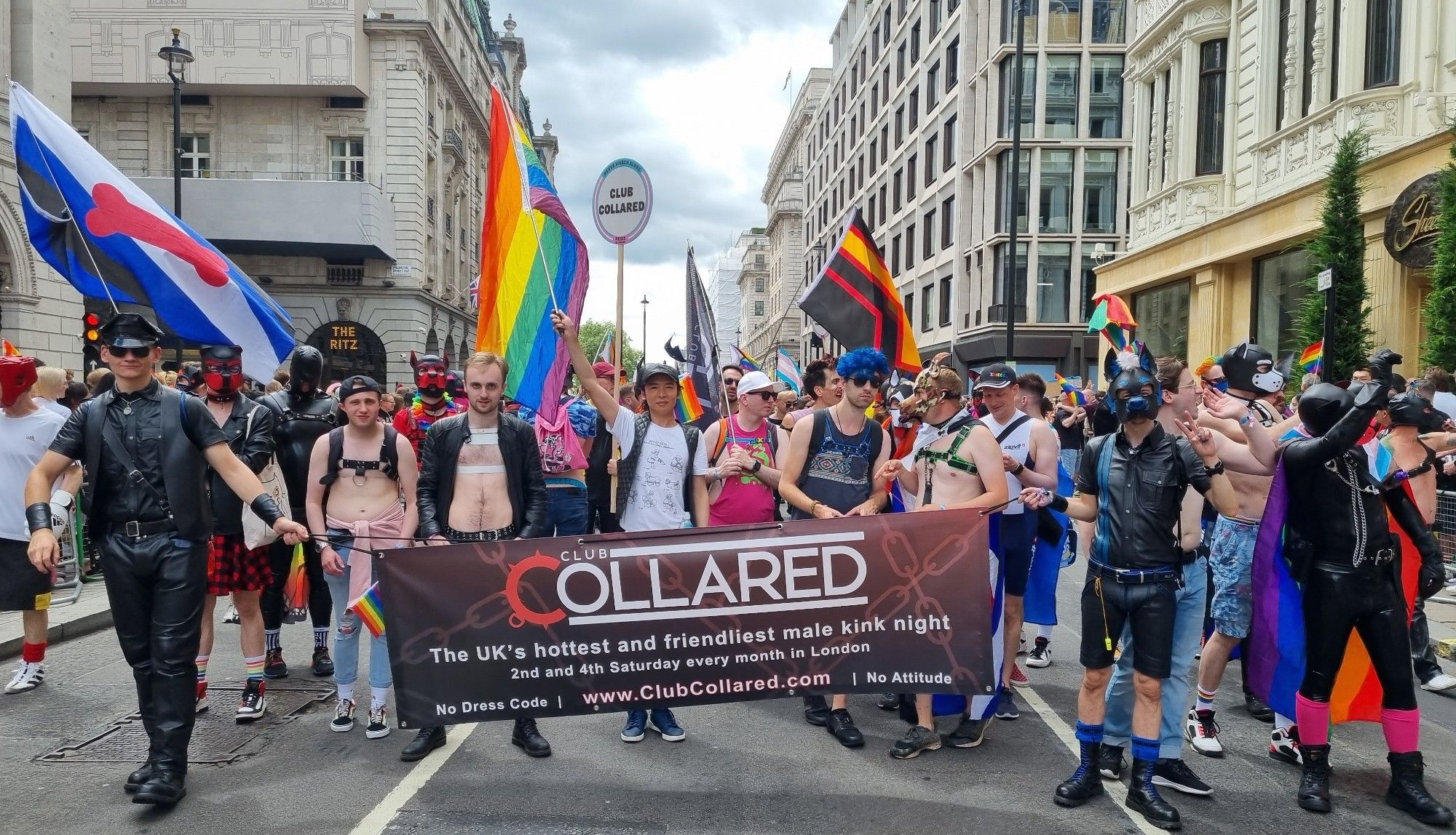 Pride in London 2023 parade group for Collared. Various kinksters behind a banner and various pride/kink flags being flown