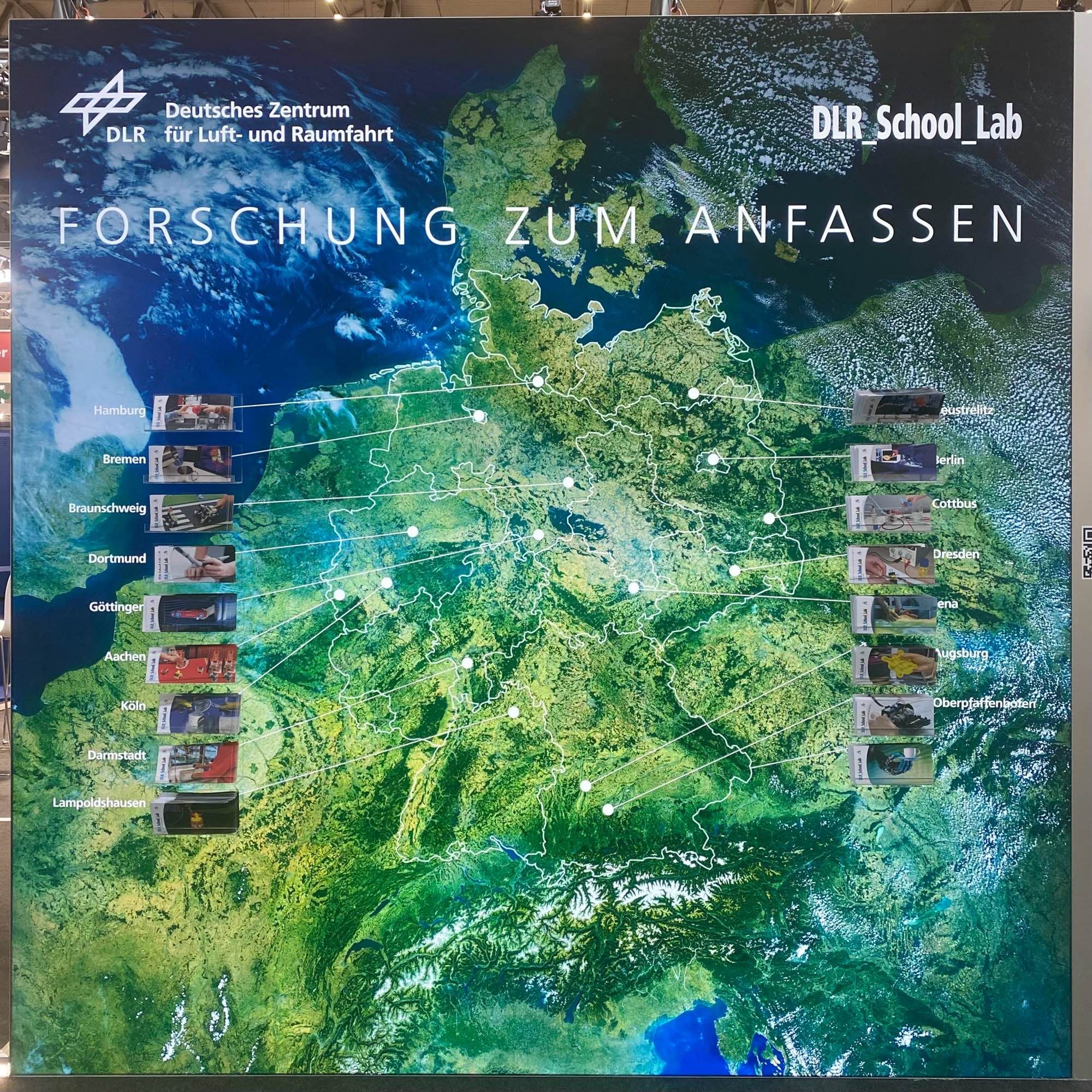 Eine Seite unseres Standes mit einem ungefähr 3 Meter mal 3 Meter großen Satellitenbild Deutschlands. Die Umrisse der Bundesländer sind mit weißen Linken markiert und an allen 16 Standorten der DLR_School_Labs sind weiße Punkte, die durch Linien mit den jeweiligen Flyer-Boxen am Rand verbunden sind. Ungefähr auf Höhe von Dänemark steht unser Slogan „Forschung zum Anfassen“.