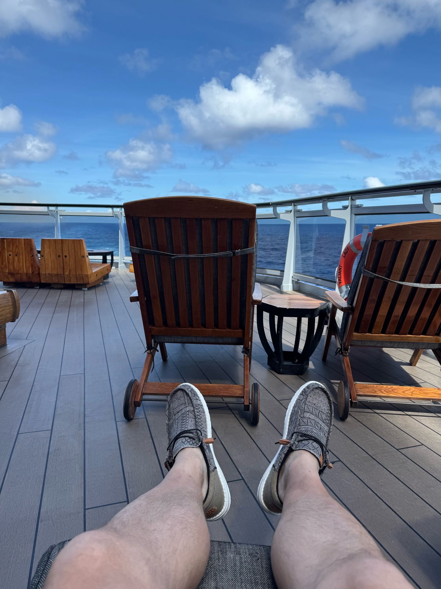 View of a deck chair and the ship's railing with blue sky and water. My legs and feet are in the bottom of the photo.