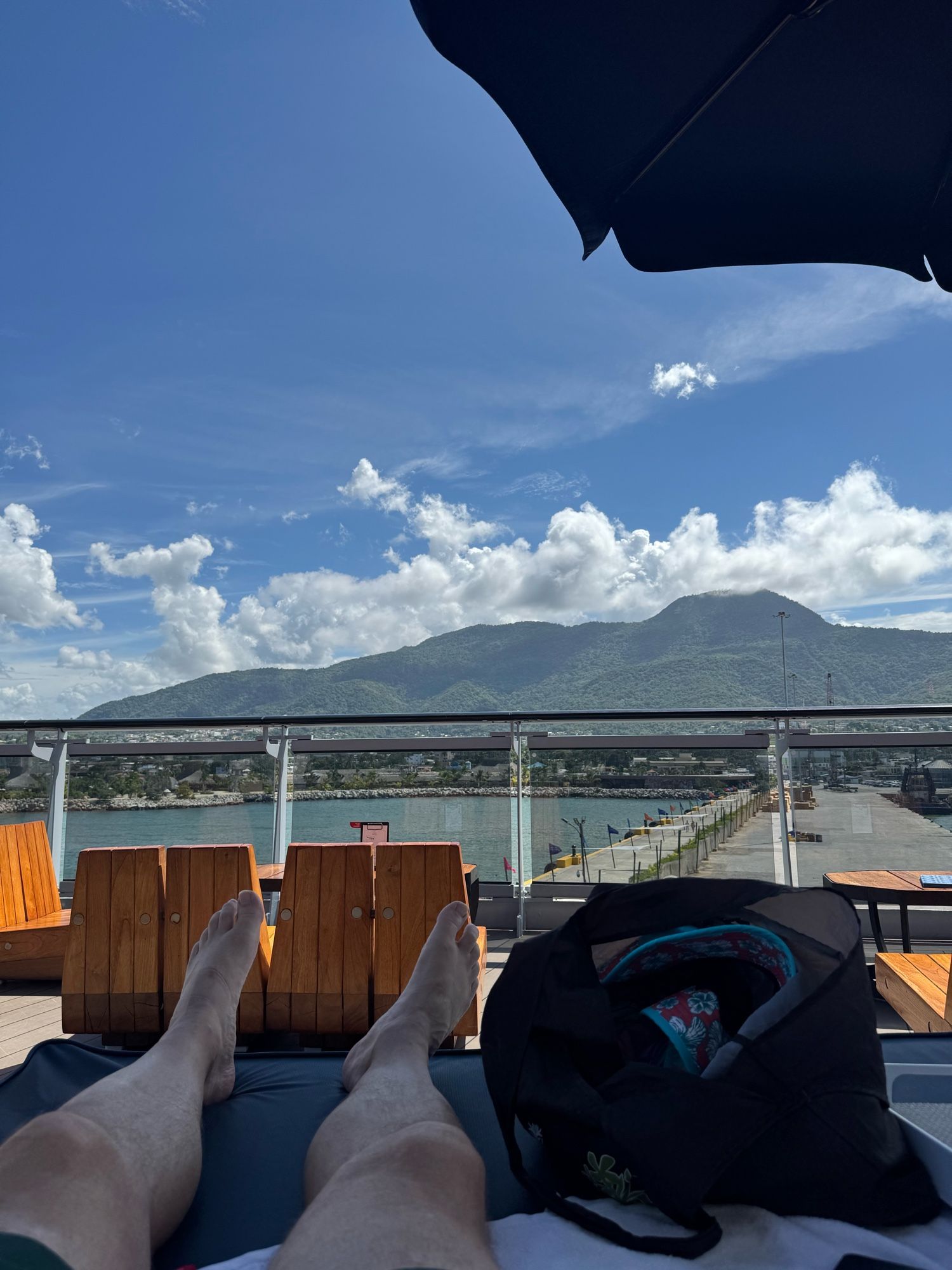 View of Puerta Plata from a daybed under sn umbrella on a cruise ship.Blue sky and white fluffy clouds over the green covered sides of Mount Isabel Torres which looms over the city.