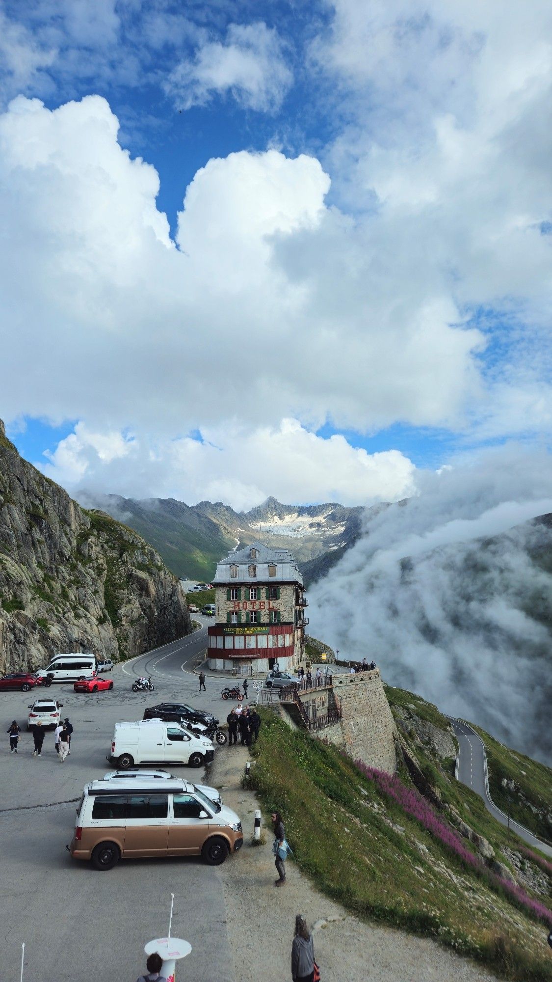 Gletscherhotel in der Lehre. Rechts geht der Berg steil herab, Blumen und Gras. Dahinter der Gletscher und Wolken die die Berge herab ziehen. Im Vordergrund Autos auf dem Parkplatz