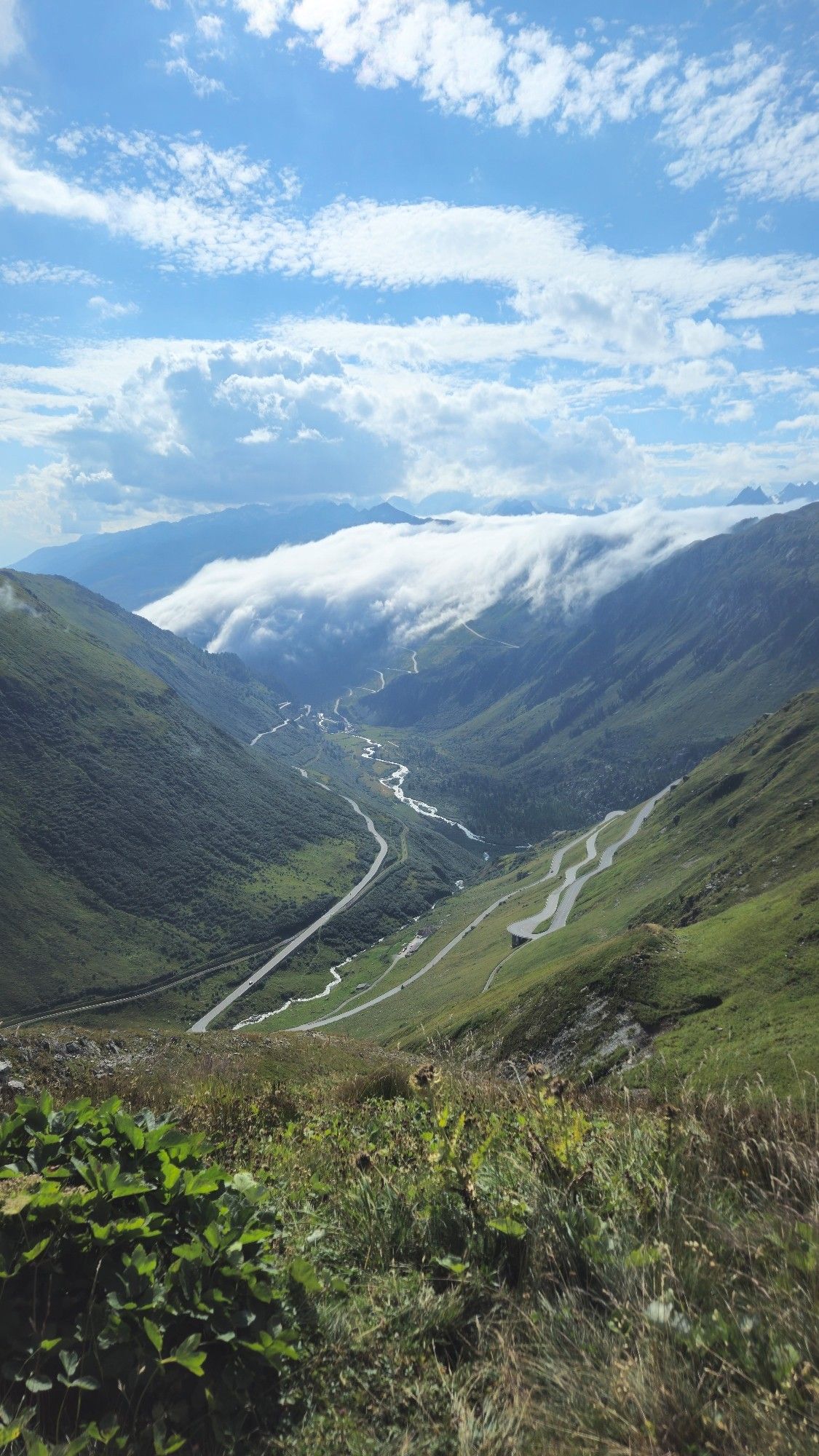 Blick vom Berg ins Tal. Von rechts schlängelt sich eine Straße den Berg ins Tal wo ein Fluss fließt. Dahinter fallen Wolken die Berge herab