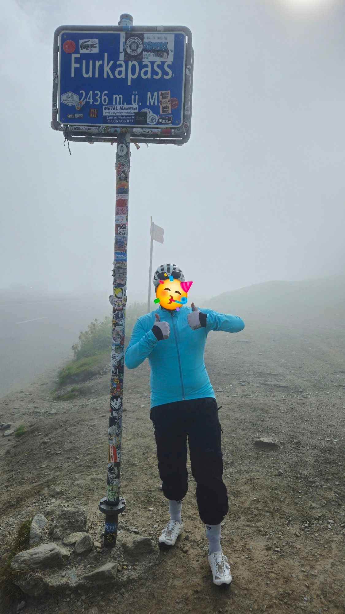 Typ mit Regenjacke und länger Hose steht am Furkapass Schild und streckt die Daumen hoch. Die Hose ist in weißen Socken gesteckt