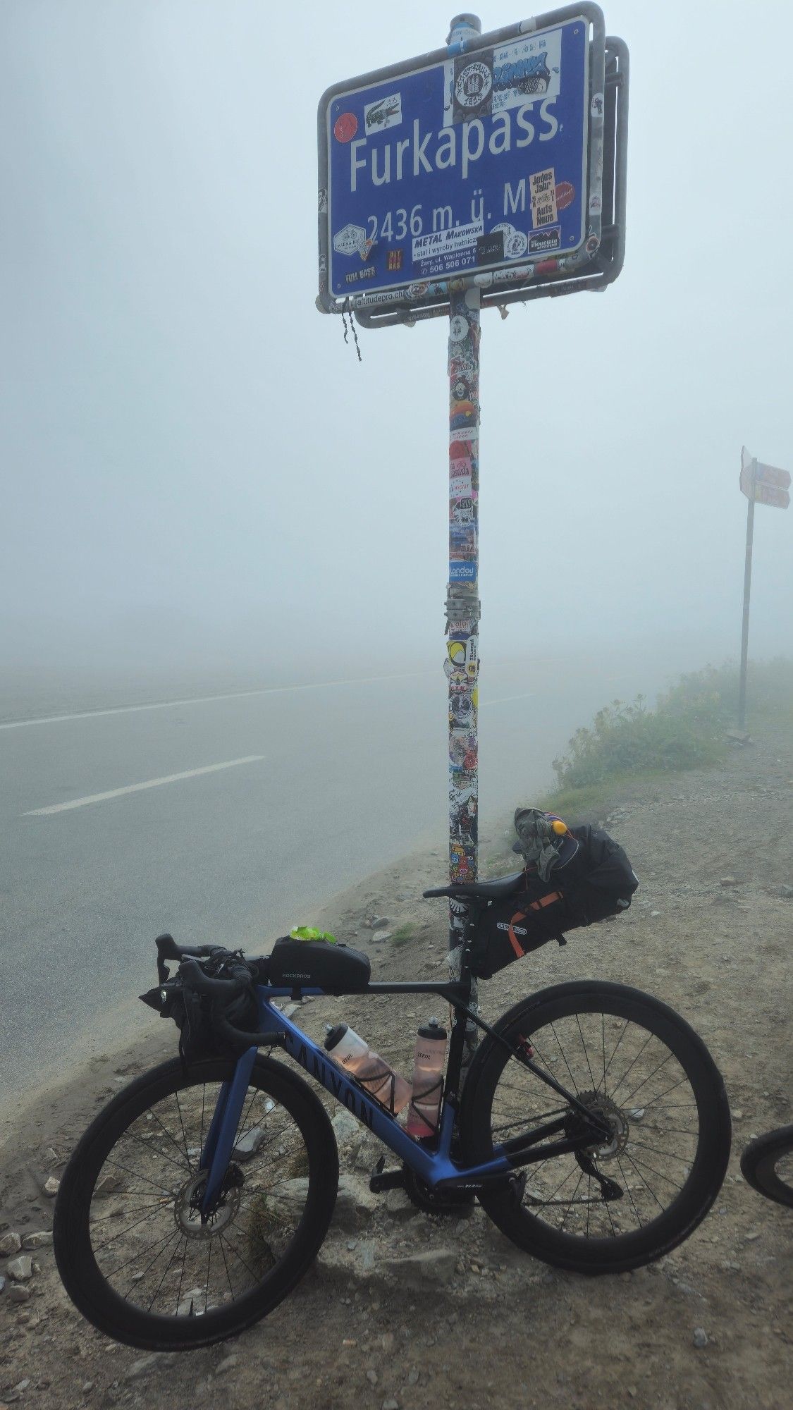 Rennrad lehnt am Furkapass Schild 2436 Meter über Null. Darunter Schotter. Die Straße und alles dahinter geht in der Wolke unter