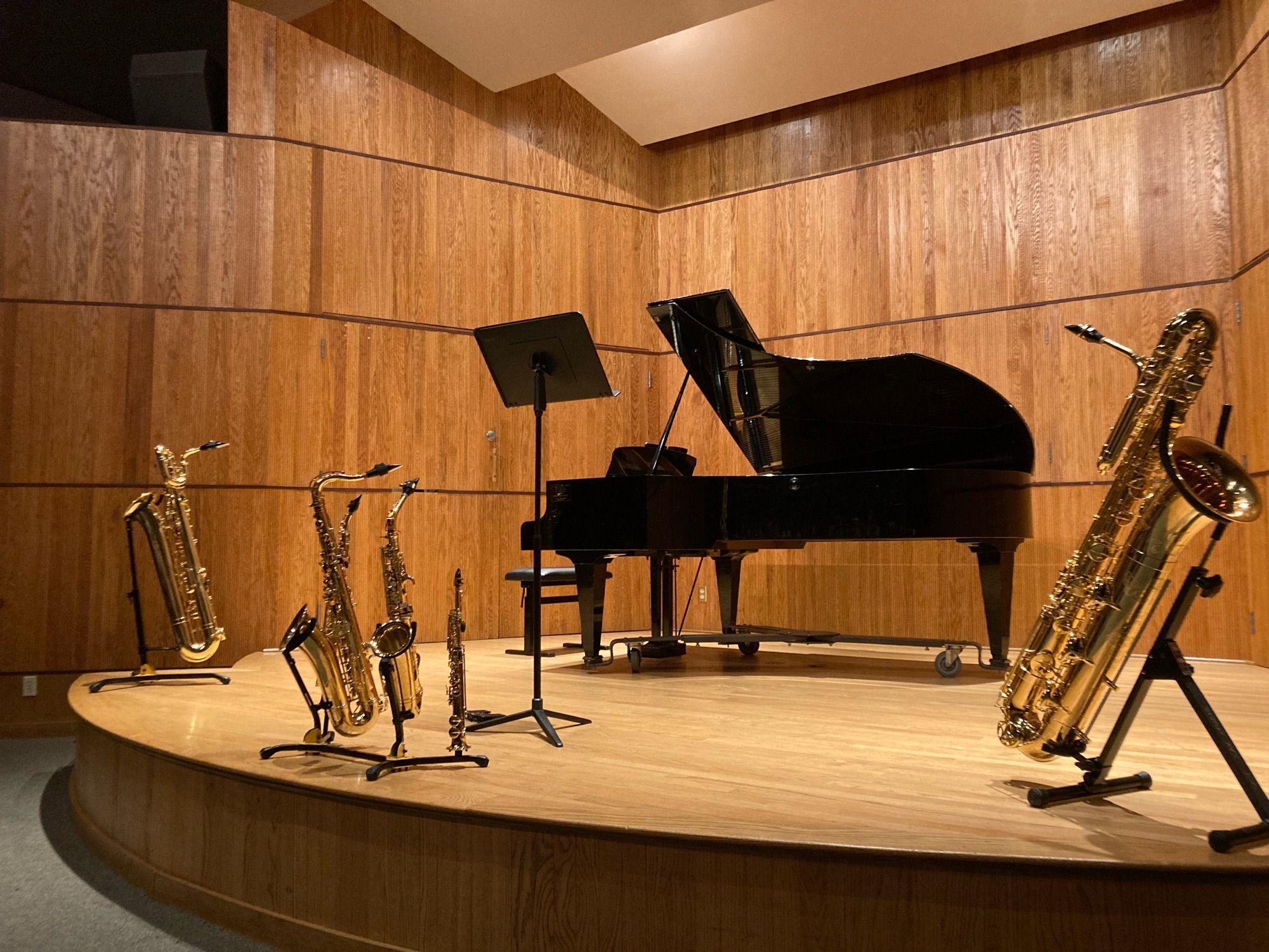 Six saxophones on a stage with a music stand and a piano. Baritone, soprano, tenor, alto, sopranino and bass.