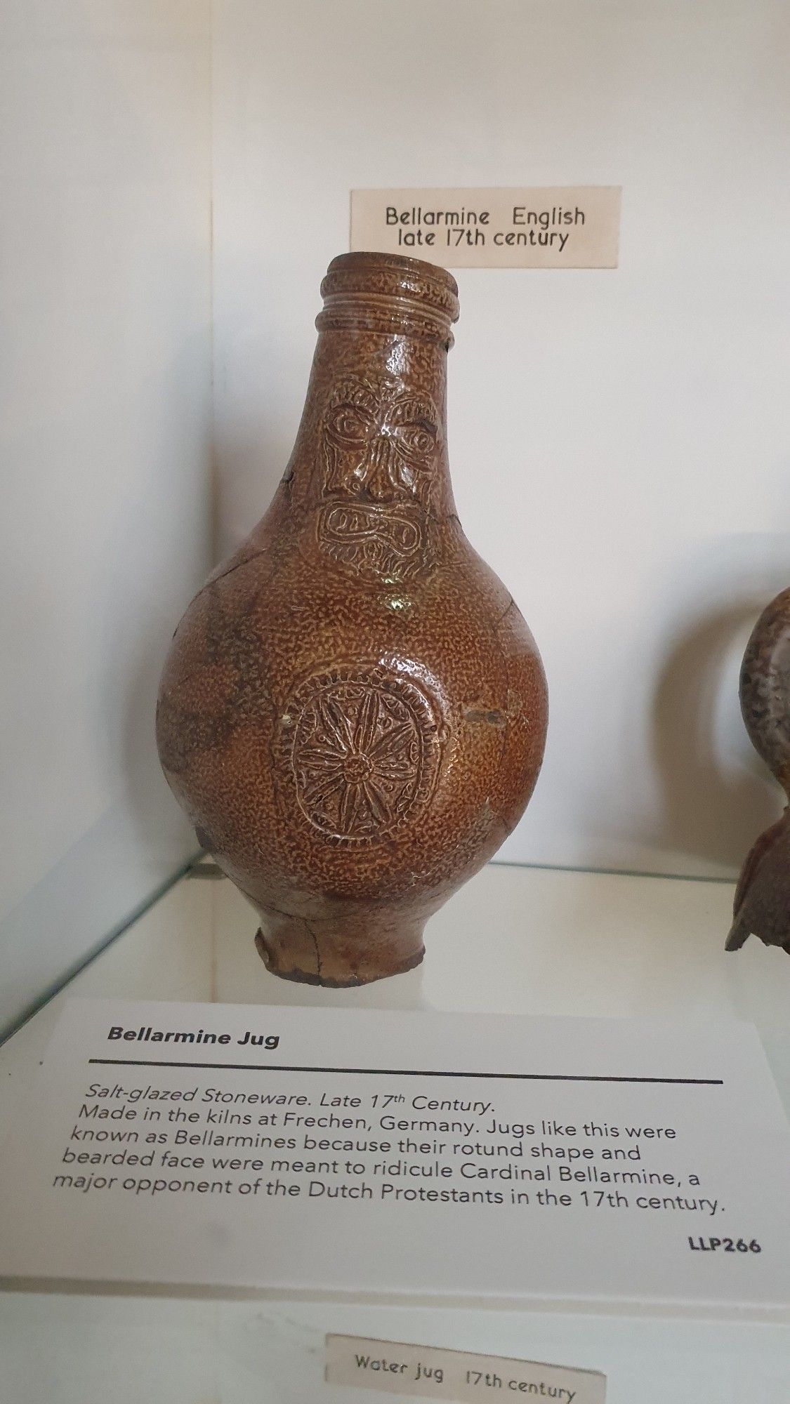 Antique jug in a museum display, labelled as: "Salt glazed stoneware jug, late 17th century. Made in the kilns at Frechen, Germany. Jugs like this were known as Bellarmine because their rotund shape and bearded face were meant to ridicule Cardinal Bellarmine, a major opponent of the Dutch Protestants in the 17th century"