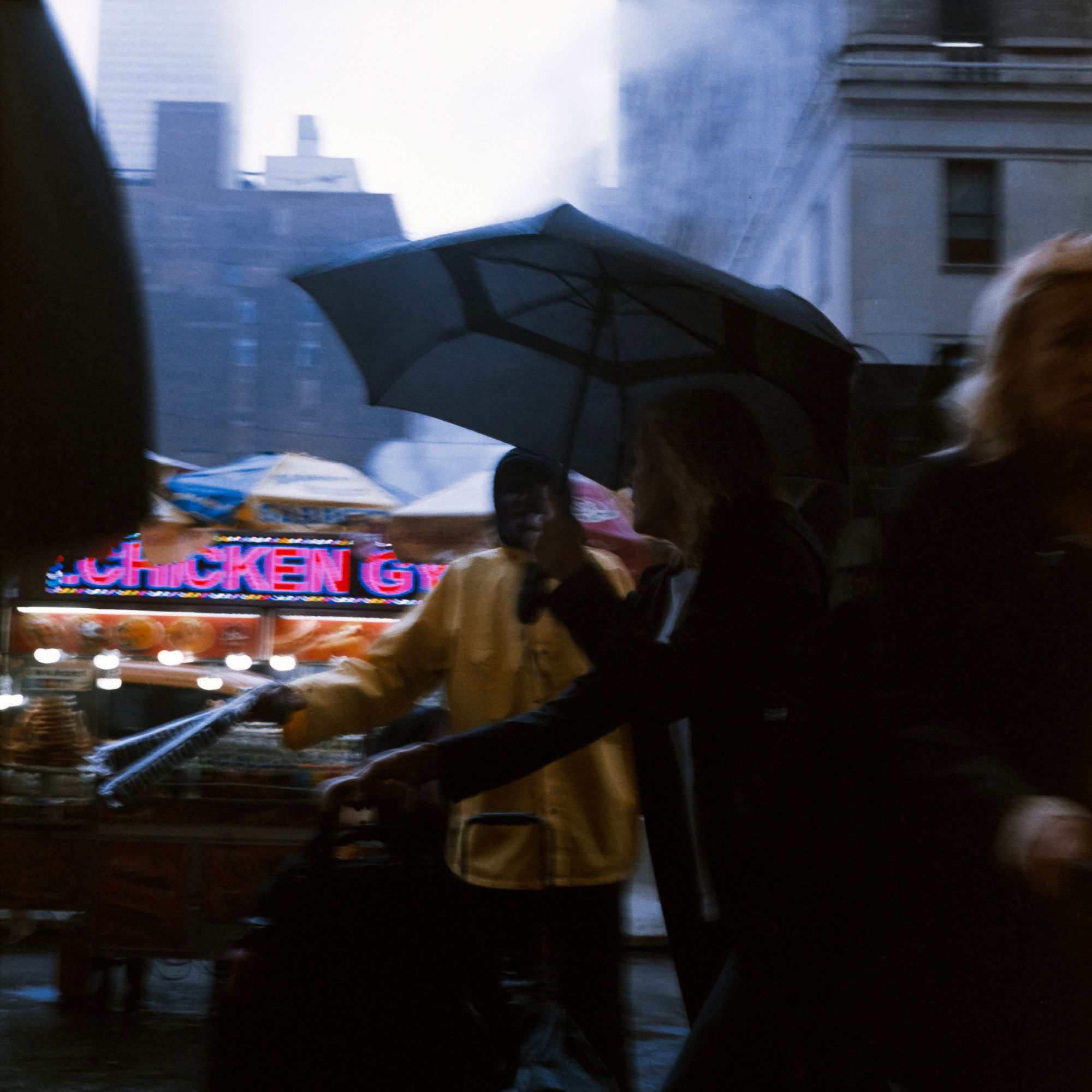 A woman in a dark jacket with an umbrella glances toward a man in a yellow raincoat as she runs past. A halal cart is in the background, its LED sign reading a neon "CHICKEN GYRO"