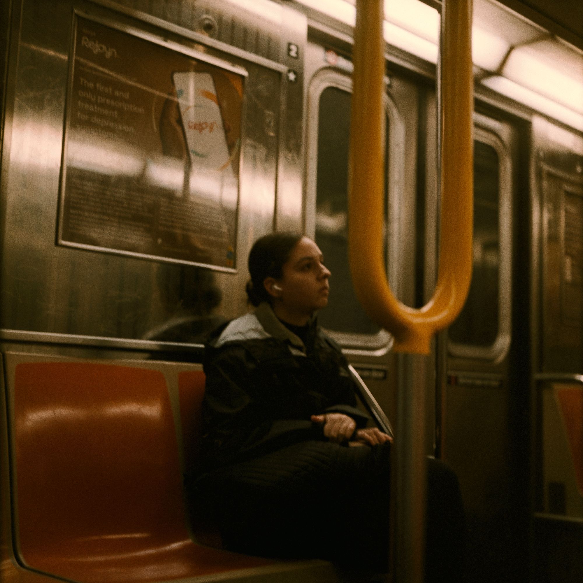 A woman sitting alone on the NYC subway, near the door to the car. She's looking away as if lost in thought.