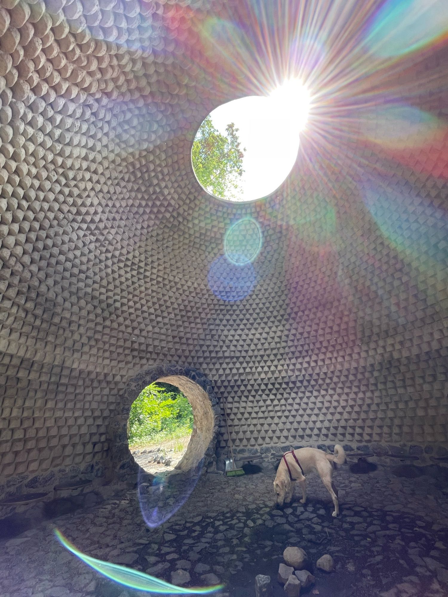 Eine begehbare Lehmkuppel mit runder Öffnung oben und als Eingang. Ein Hund steht am Rande einer Vertiefung in der Mitte der Kuppel. Durch die obere Öffnung scheint die Sonne und ein buntes Lichtspiel verteilt sich in der Kuppel.