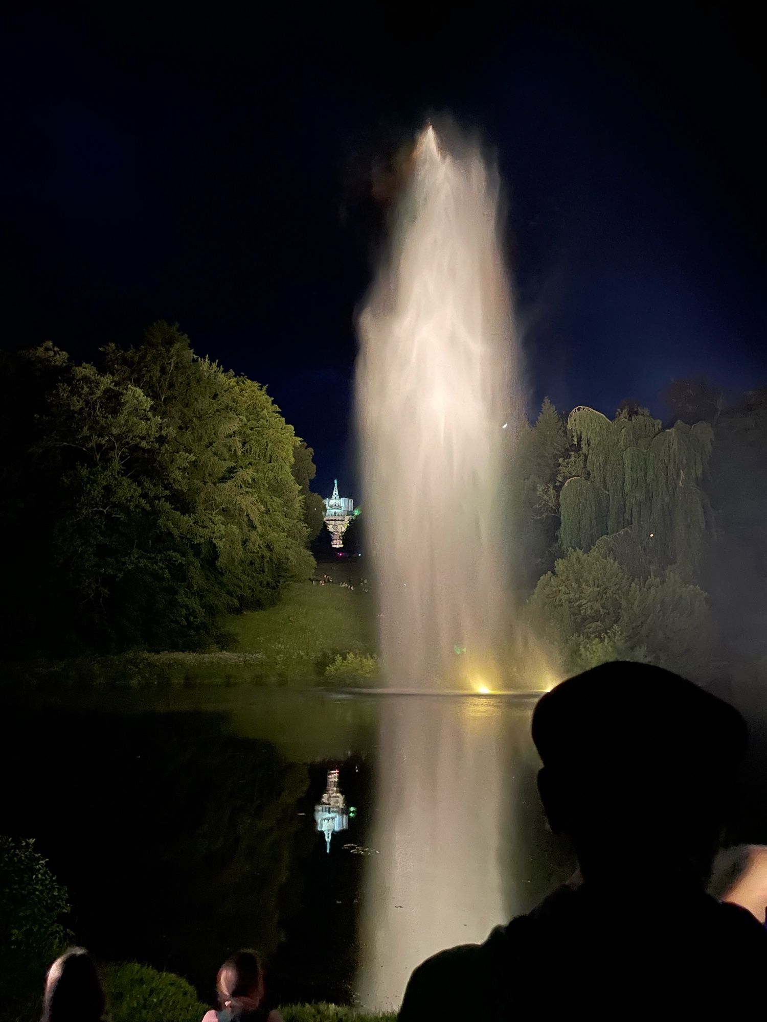 Dunkler Nachthimmel, mittig eine hohe Fontäne, links davon der beleuchtete Herkules mit Kaskaden, die sich im Fontänenteich spiegeln. Rechts und links von der Fontäne Bäume im Hintergrund. Hinterköpfe im Vordergrund.