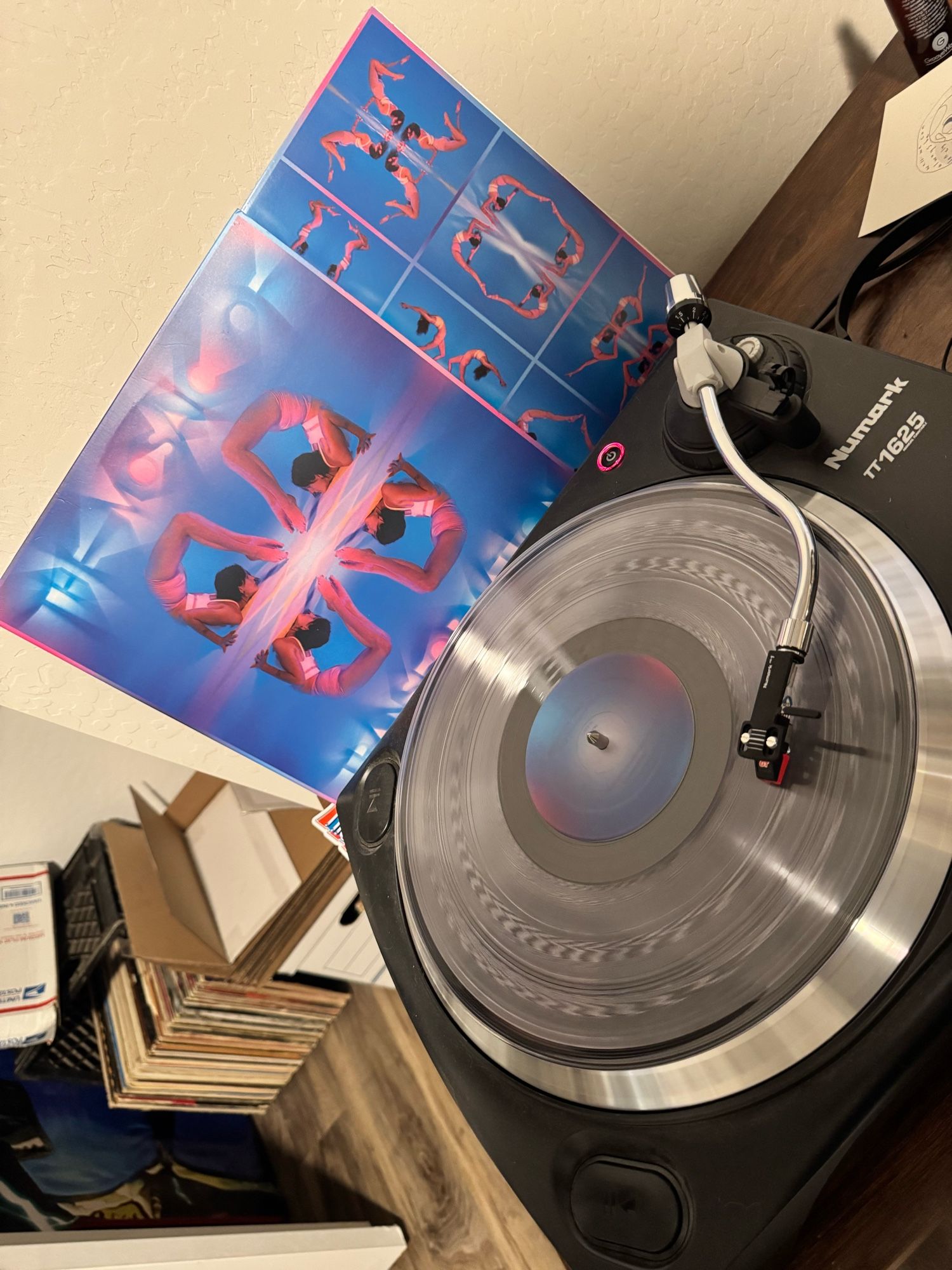 The mosaiac transformation by Kaitlyn Aurelia Smith, transparent record spinning on a turntable. Cover and inner sleeve propped up next to it. Cover and sleeve look like a kaleidoscope effect of a woman in a scorpion like pose over a blue pink-lit background