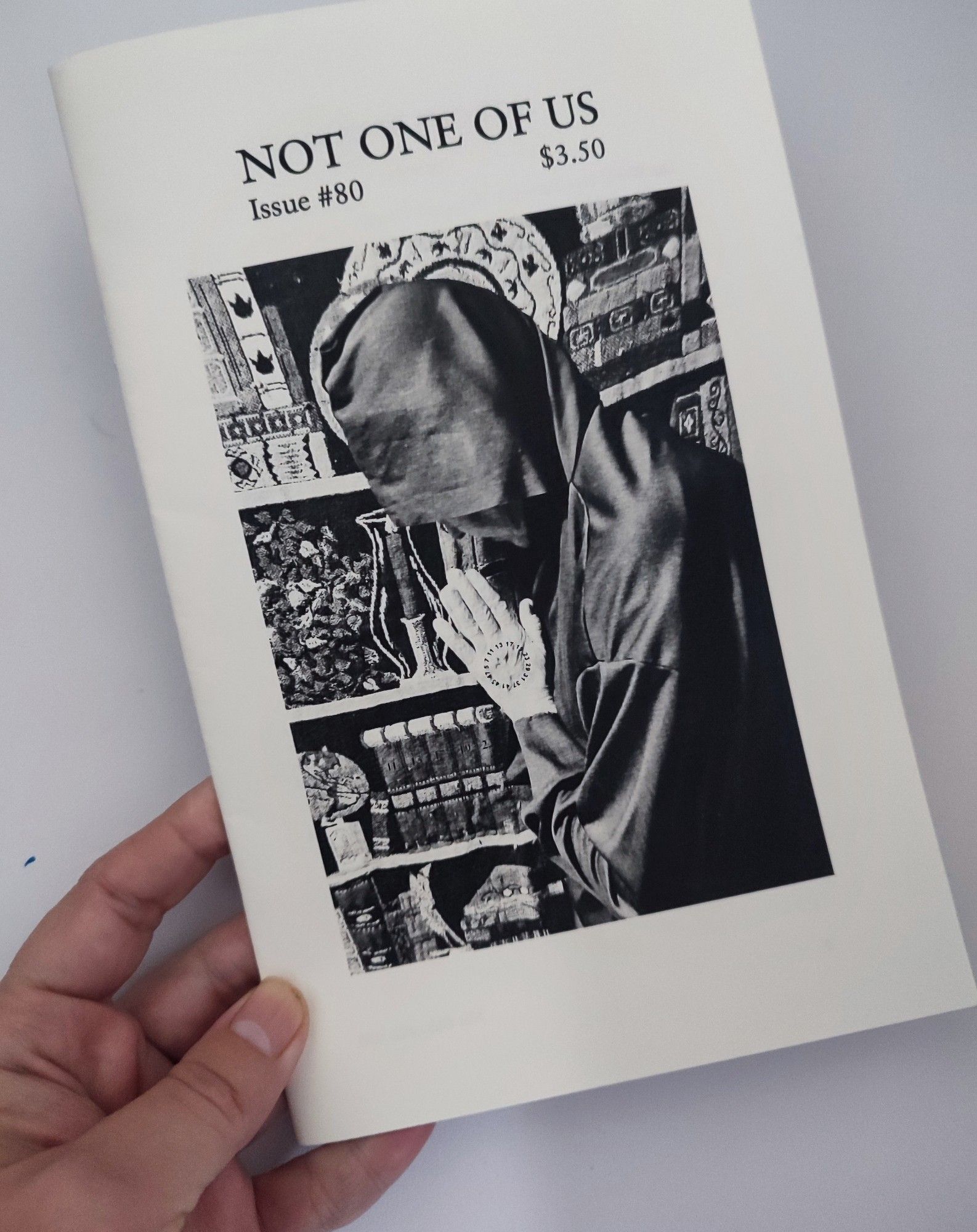 A hand holding a black and white printed zine. A hooded figure with strange markings prays before a tapestry. The title is printed in the top white border. Not One Of US, Issue #80, $3.50