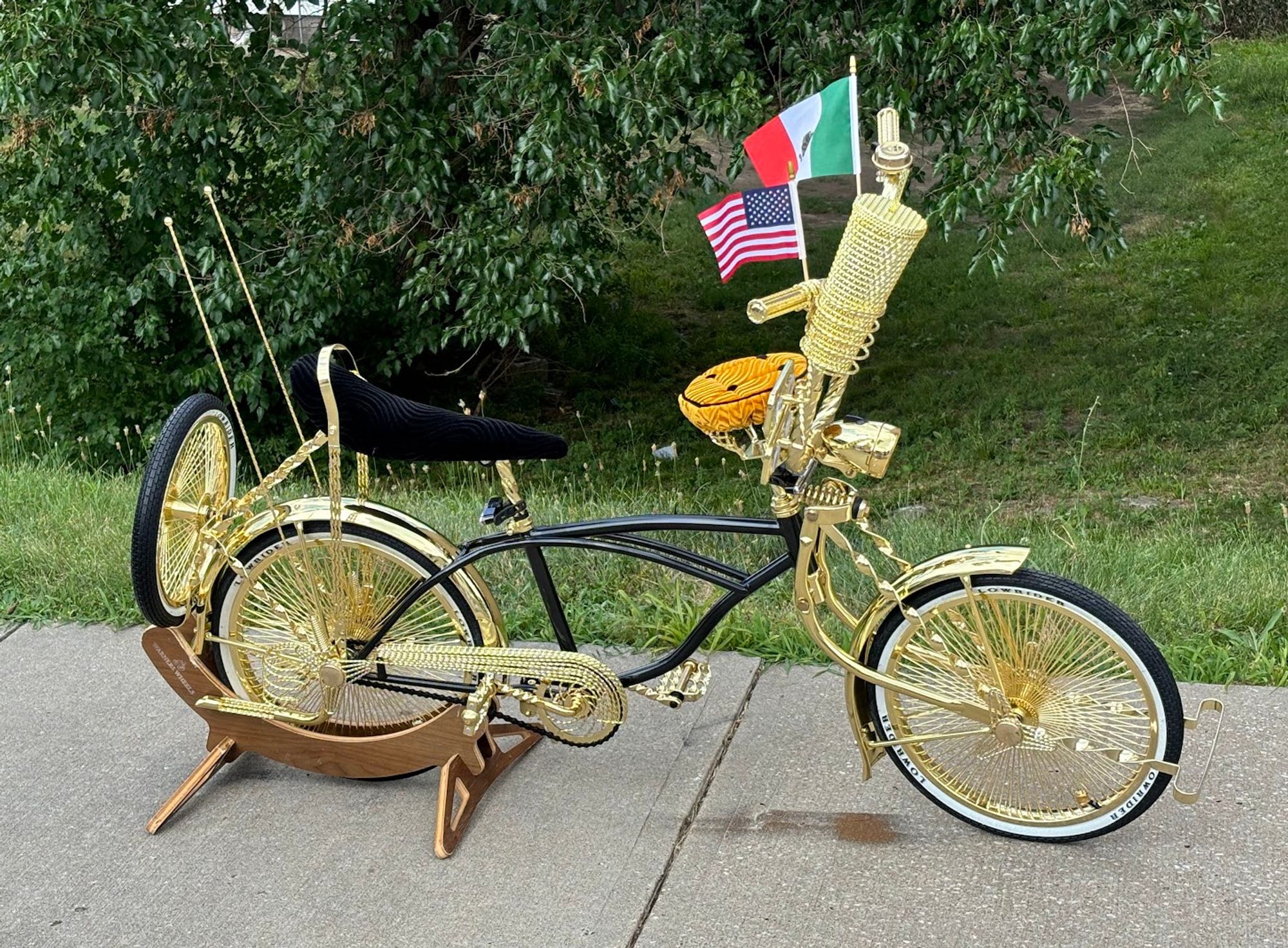 this is another angle of the previous, triple-gold detailed bike ,showing a couple more details.  the bike is leaning on a wooden rear-wheel stand on a sidewalk, in front of green grass.