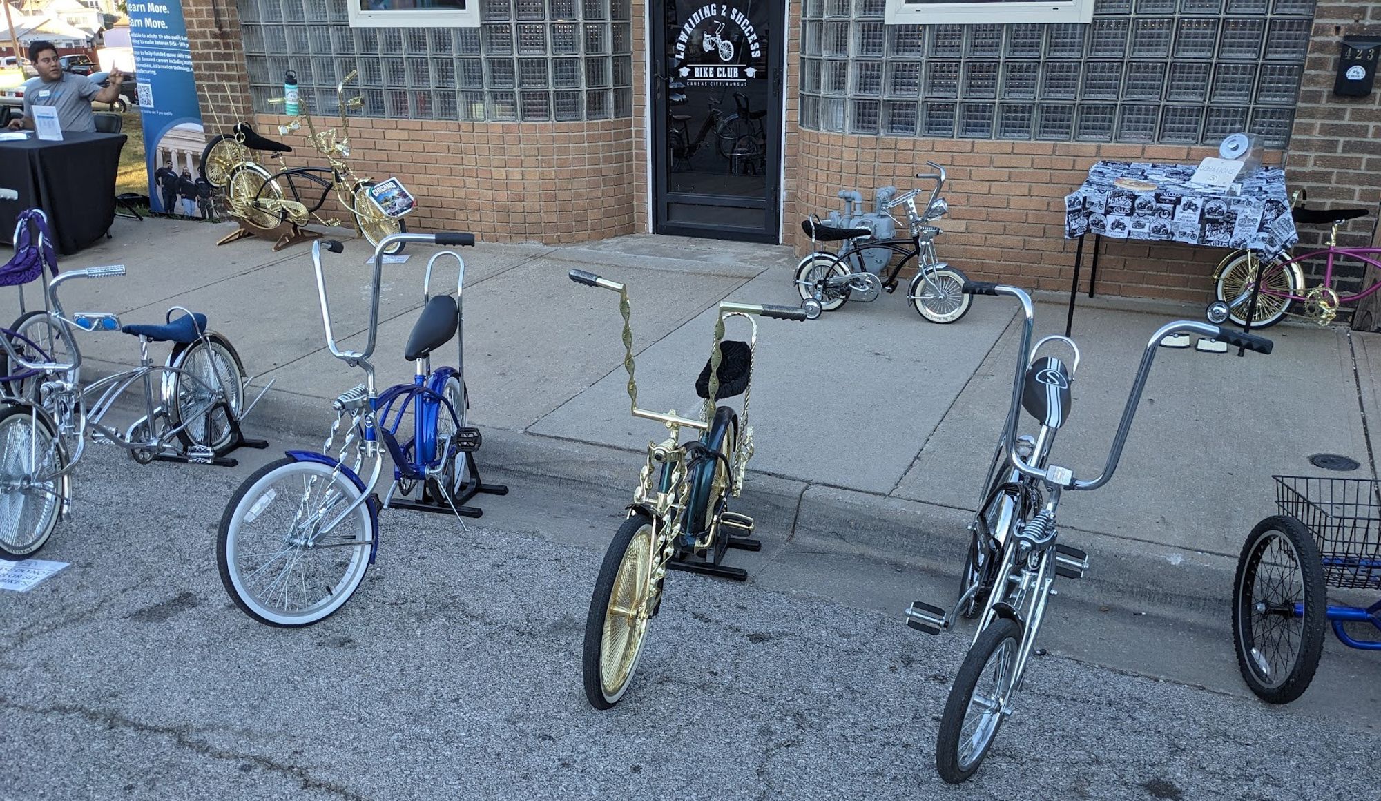 picture taken on the street in Kansas City Kansas, of the Lowriding 2 Success Bike Club, with at least 9 bikes visible.  All the bikes are custom lowriders, with one tricycle.  two of the bikes are gold detailed, and the rest are chrome.