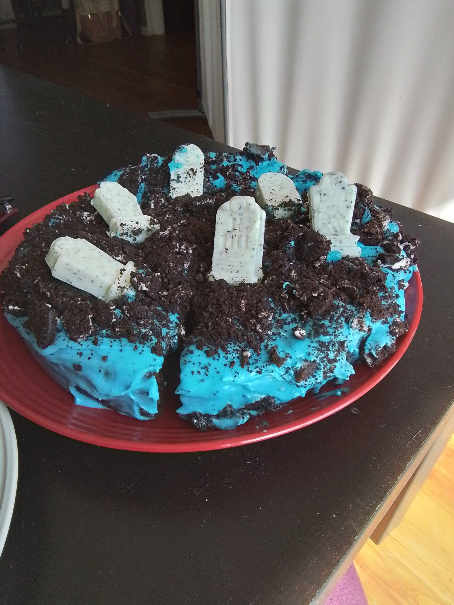 The top layer of the pumpkin chocolate cake with blue cream cheese icing is pieced together on a red plate. It had fallen apart and was reassembled. It has oreo-like cookie crumbles on top and candy tombstones.