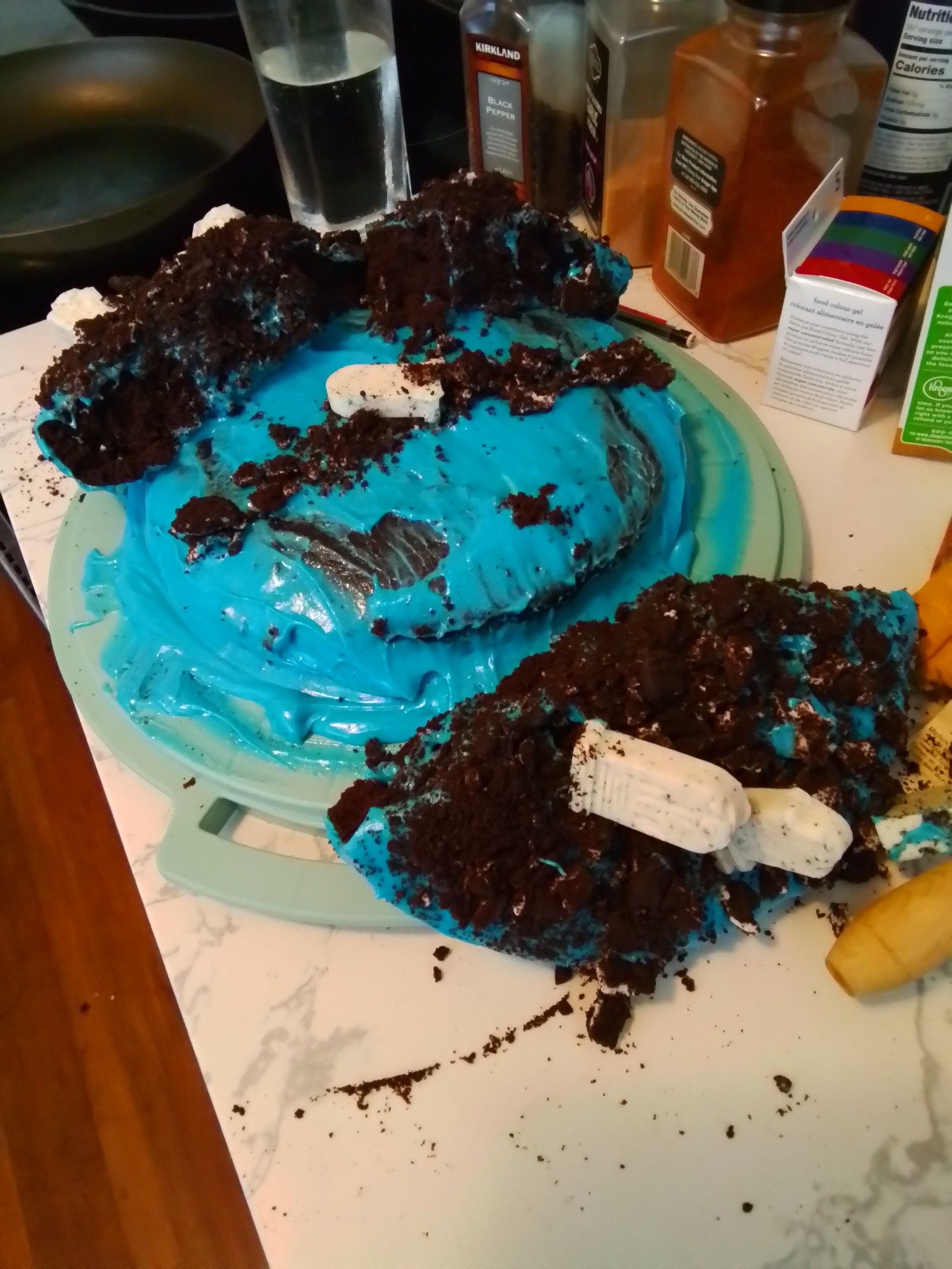 A once two-tiered chocolate pumpkin cake with blue icing has its the top layer ripped in half. The cookie crumble on top with candy tombstones is everywhere.