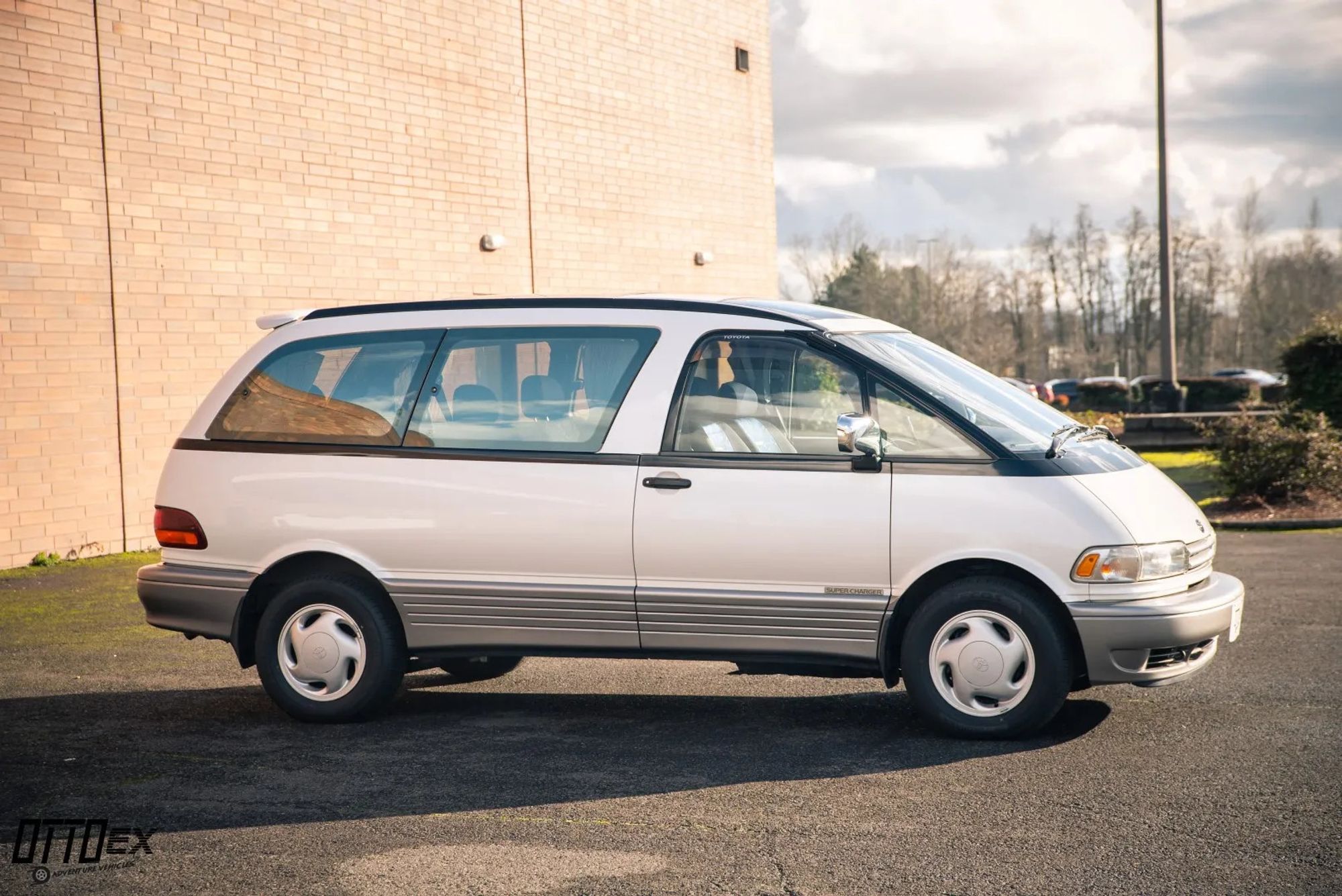 A white 1994 Toyota Previa minivan parked in a parking lot. The van has a silver stripe along its side and alloy wheels.