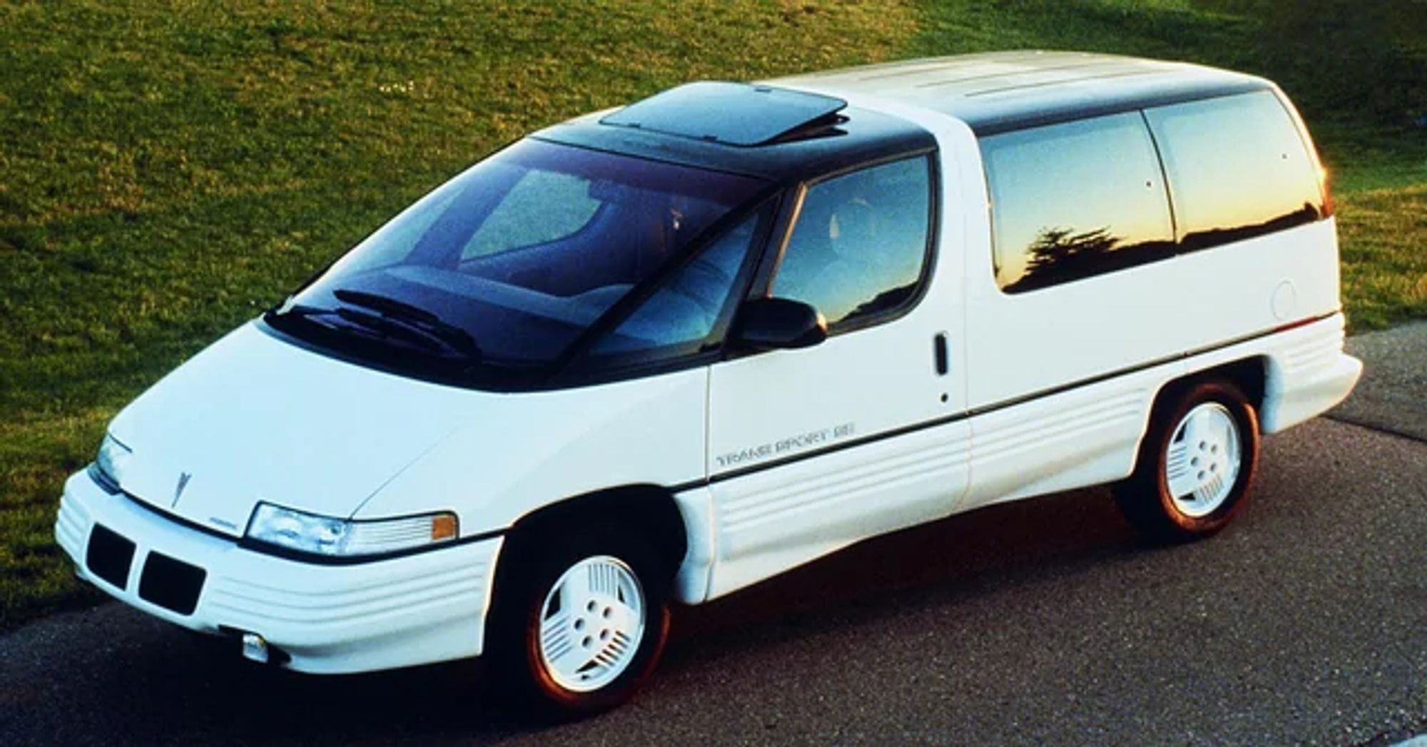 A white 1990 Pontiac Trans Sport minivan parked on a grassy hill. The van has a unique design with a black roof and a large front windshield.