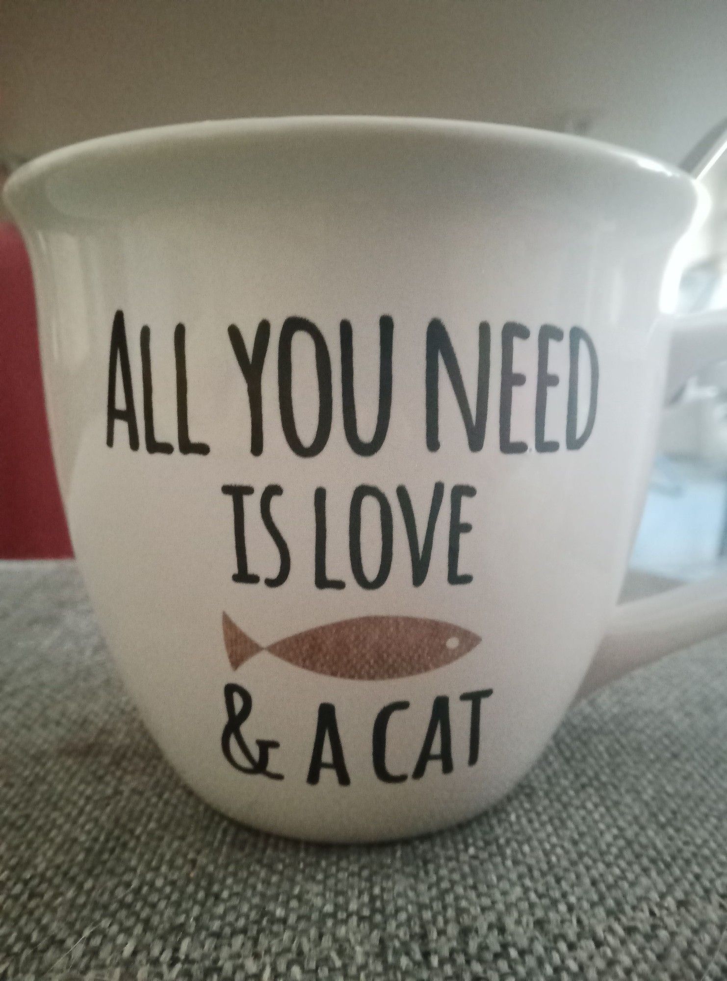 eine weiße Kaffeetasse mit der Aufschrift "All you need is love and a cat" und einem goldenen Fisch