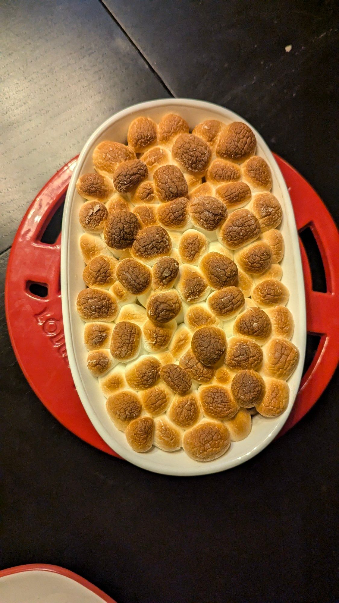 A small baking dish on a hot plate on a table. Inside the dish is puffed-up and browned marshmallows, under which is melted milk chocolate and white chocolate. This is s'mores dip