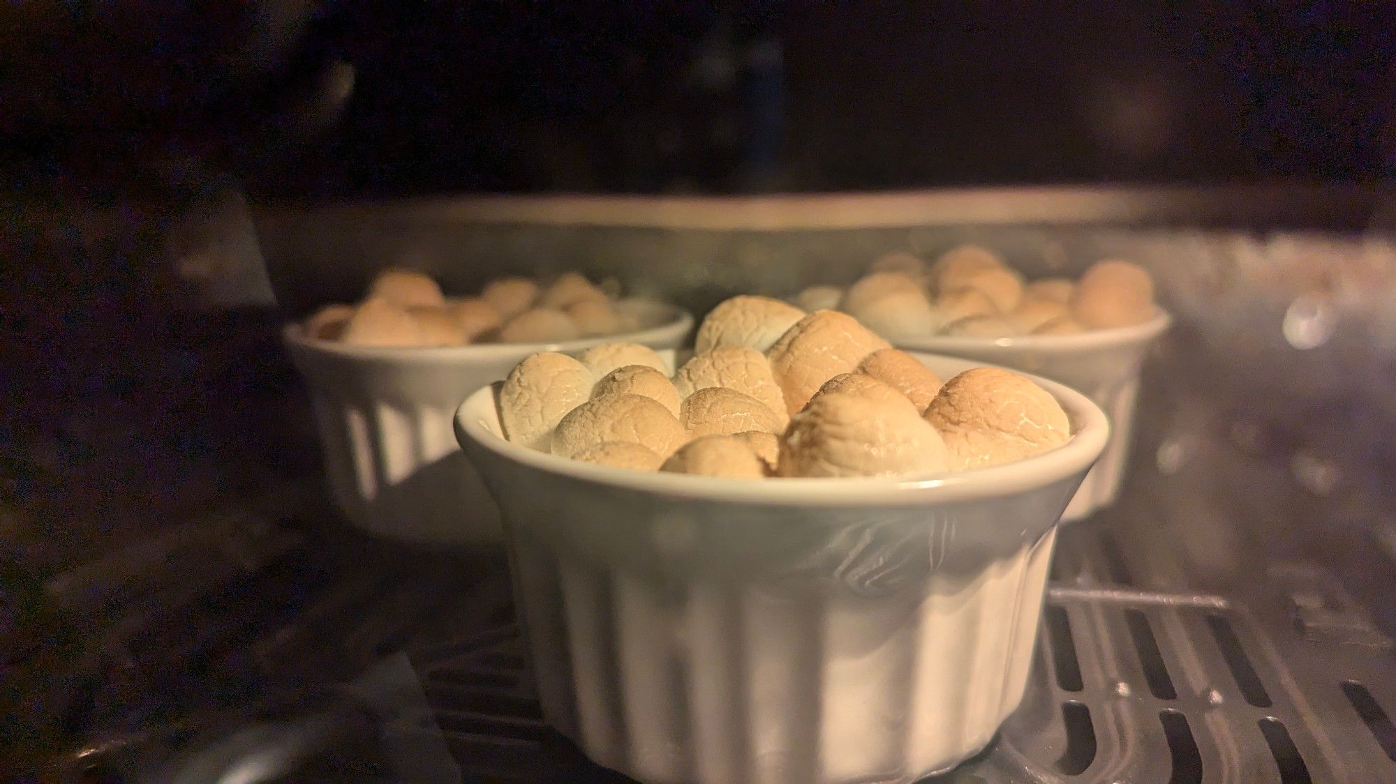 A picture of the inside of an air fryer with a window, showing three ramekins filled with marshmallows puffing up and browning in the heat