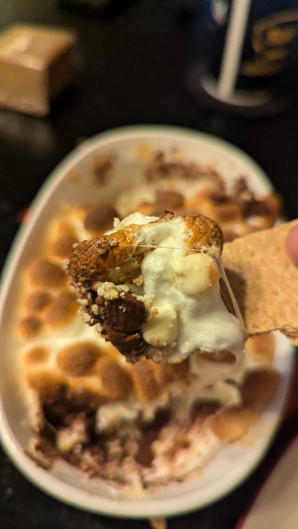 A close-up picture of the contents of the s'mores dip, scooped up with a graham cracker. Behind the close-up is the same dish of s'mores dip from the prior picture