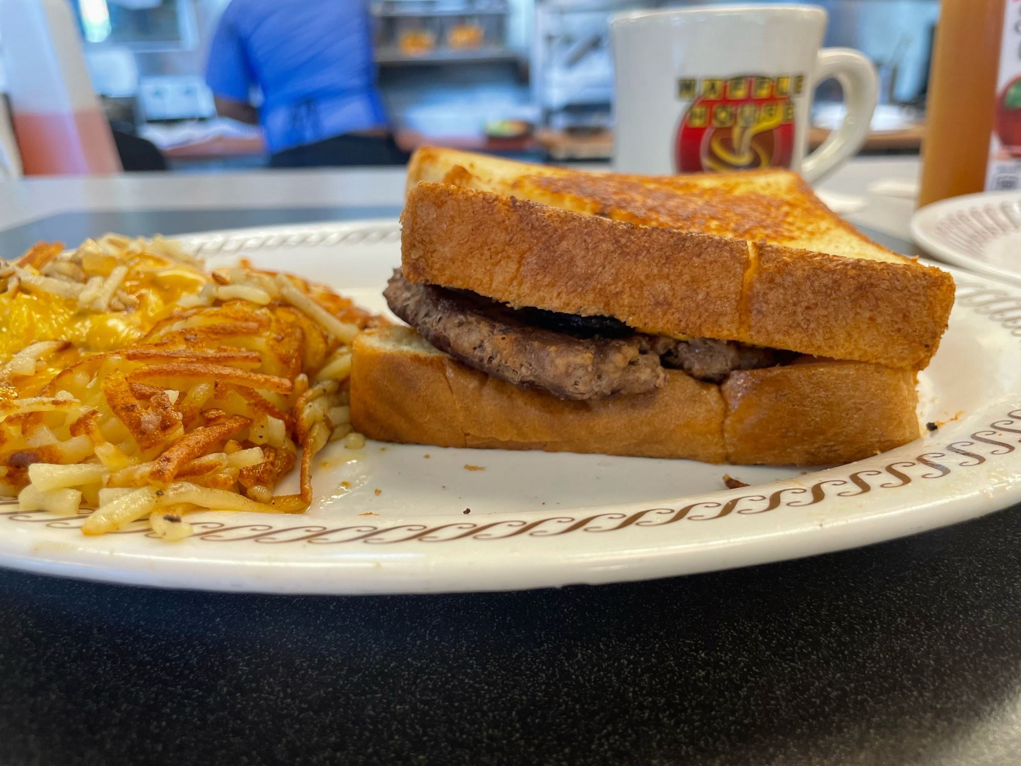 On the counter, a plate of scattered smothered capped and covered hashbrowns, patty melt, and Waffle House-branded coffee mug