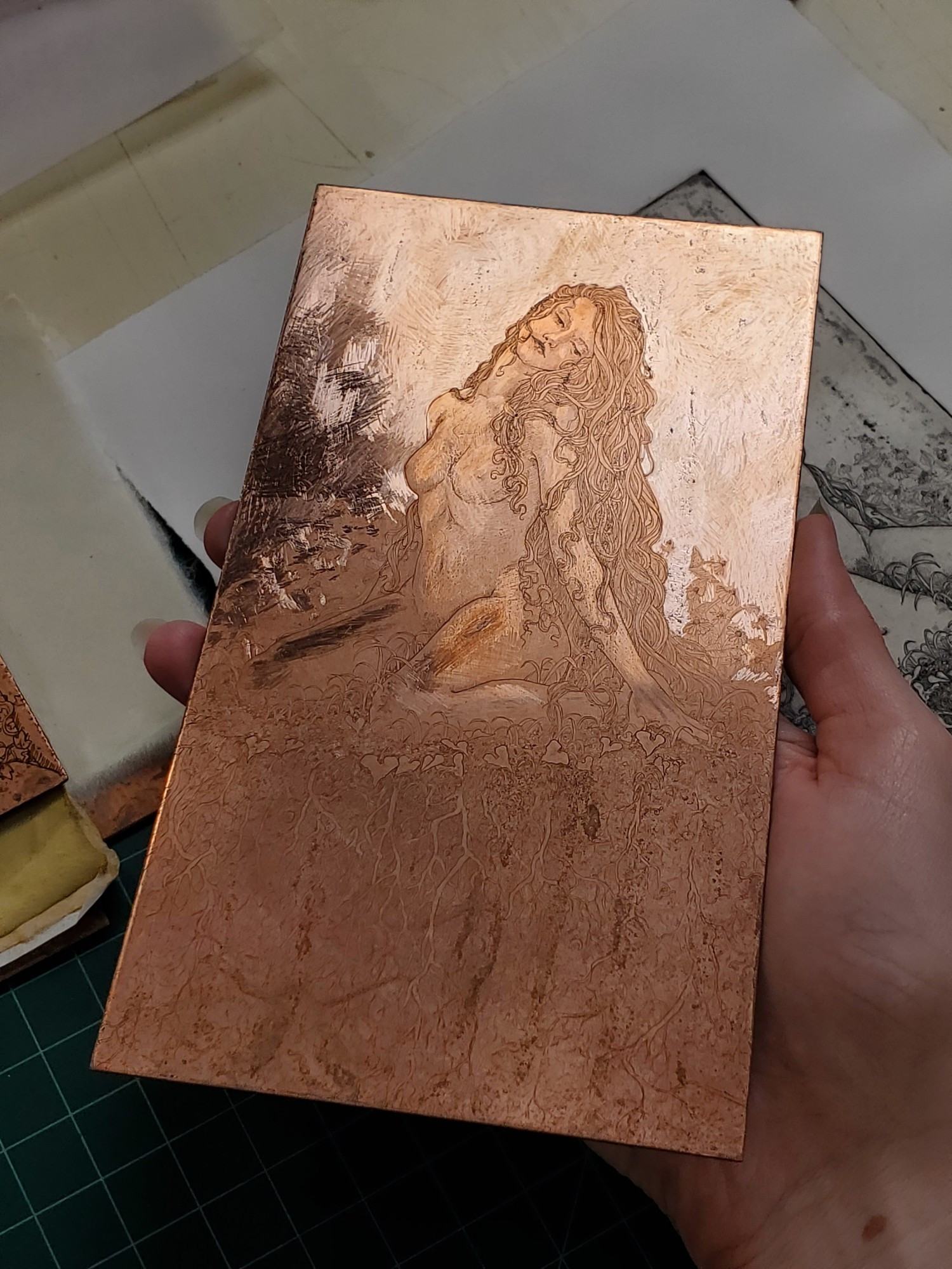 Photo of a hand holding a 4 by 7 inch copper plate. Light hits the top half of the plate, revealing the image etched into the copper: a seated female figure with long flowing hair.