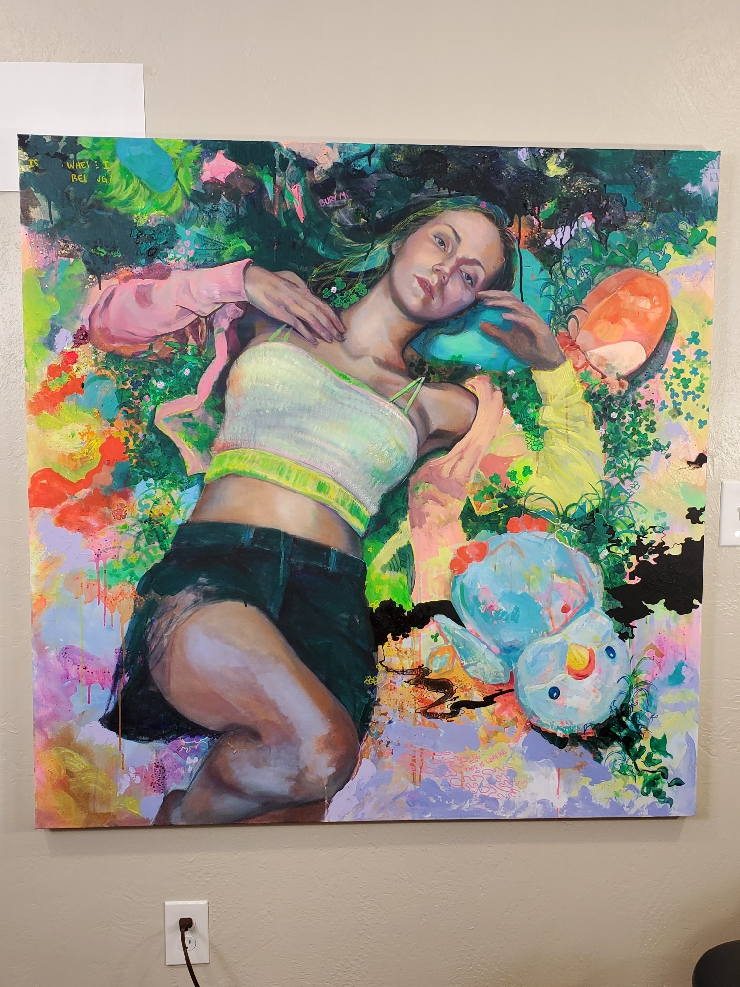 Photo of a 48x48" acrylic painting on canvas. It depicts an overhead view of a young woman laying down wearing a yellow/pink cardigan, a white crop top, and dark green shorts. The surroundings are mostly abstract but contain a few stuffed animals and foliage ground cover. The color pallette is vibrant with some areas of darkness.