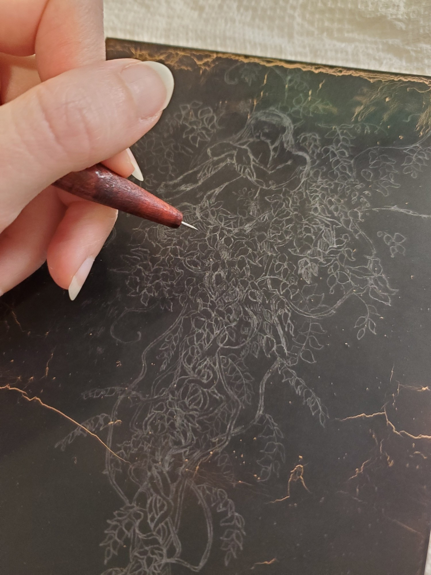 Photo of a copper plate coated with black hard ground. A graphite transferred image is on the hard ground: the preliminary drawing for "Held by a Thread." A hand is poised over the plate holding a needle point etching tool.