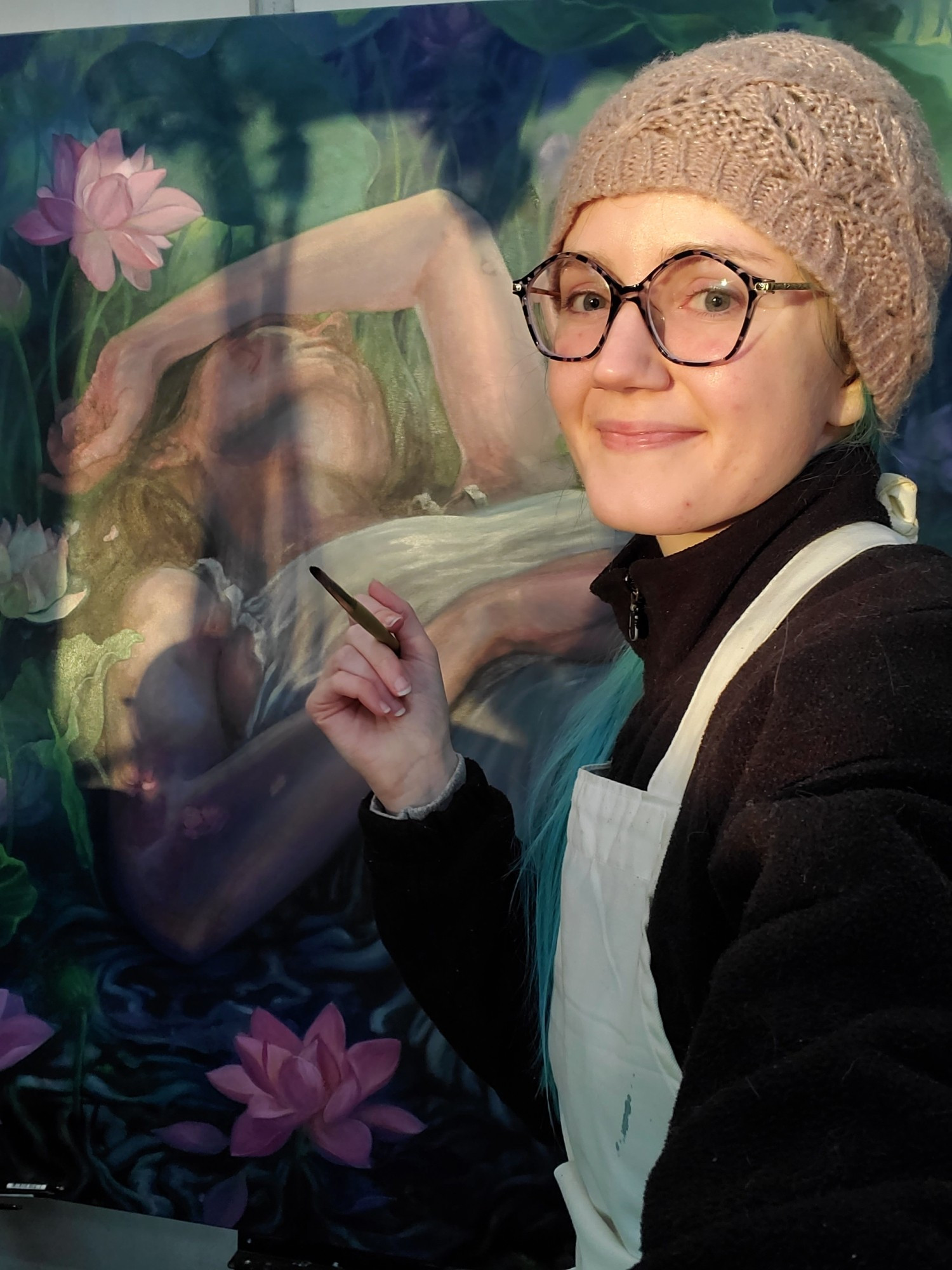 Selfie holding a paint brush in front of a large oil painting. I am wearing black rimmed glasses, a mauve knitted hat, black fleece jacket, and a white apron. I'm smiling and there is warm evening sunlight.