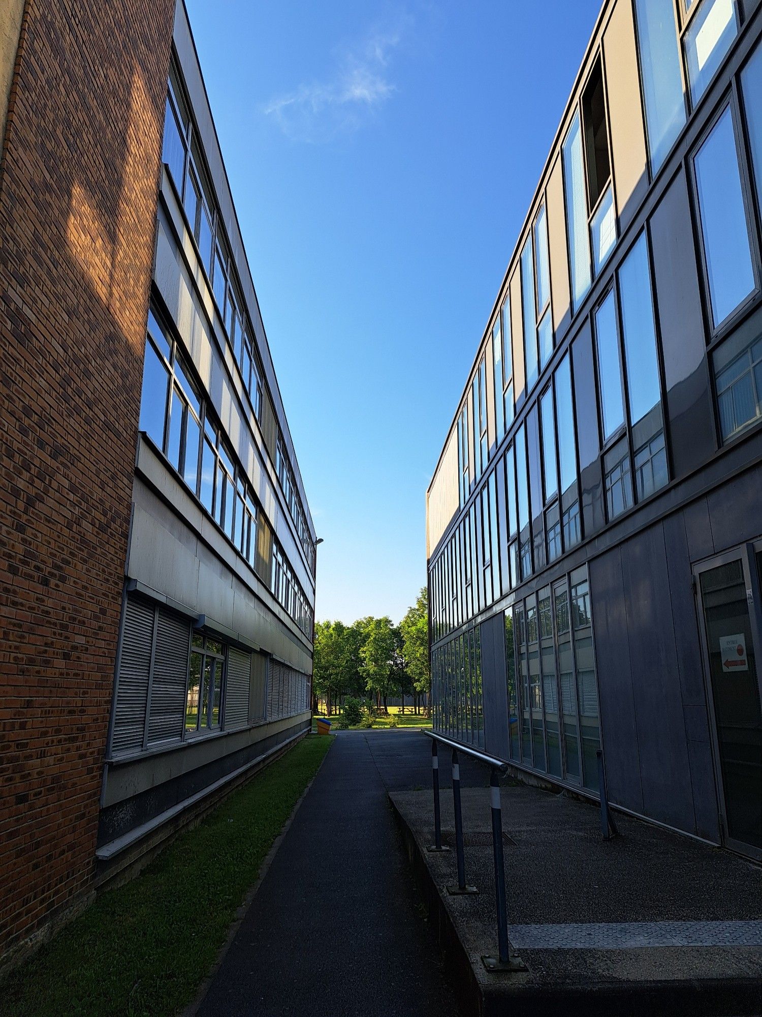 Allée en bitume entre 2 bâtiments
A gauche, en brique et petites fenêtres 
A droite verre et métal 
Le ciel est bleu, la perspective emmène l'œil vers le point de fuite dans lequel on voit des arbres.
Le haut de la photo est lumineux, ensoleillé, le bas est dans l'ombre