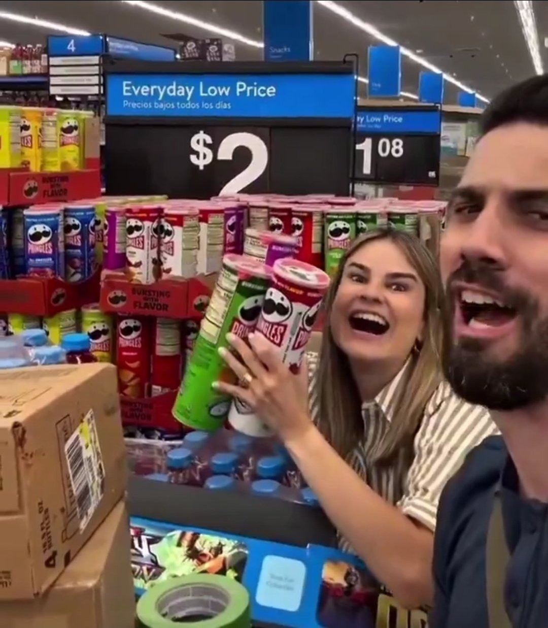 Um homem e uma mulher olhando para a câmera, em um supermercado americano. A mulher segura 3 embalagens da batata pringles nas mãos e exibe um sorriso de boca aberta. Atrás dela está a gôndola das batatas vendidas em promoção por 2 dólares americanos.  A mulher tem uma aliança do tamanho de um bracelete no dedo anelar da mão esquerda. 