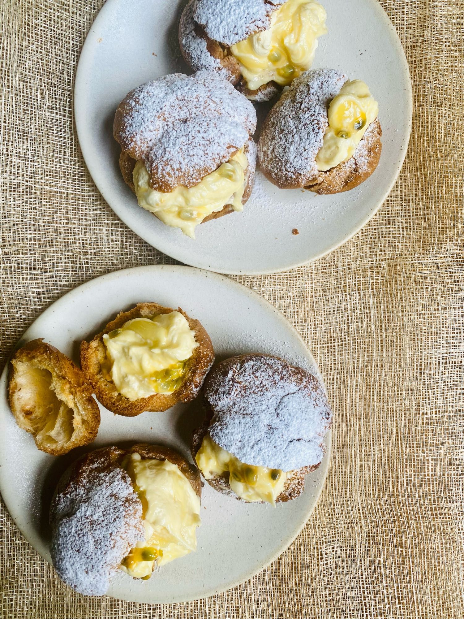 Two plates of choux buns showing their filling of lemon, cream and passionfruit. They sit on a stone coloured linen cloth.