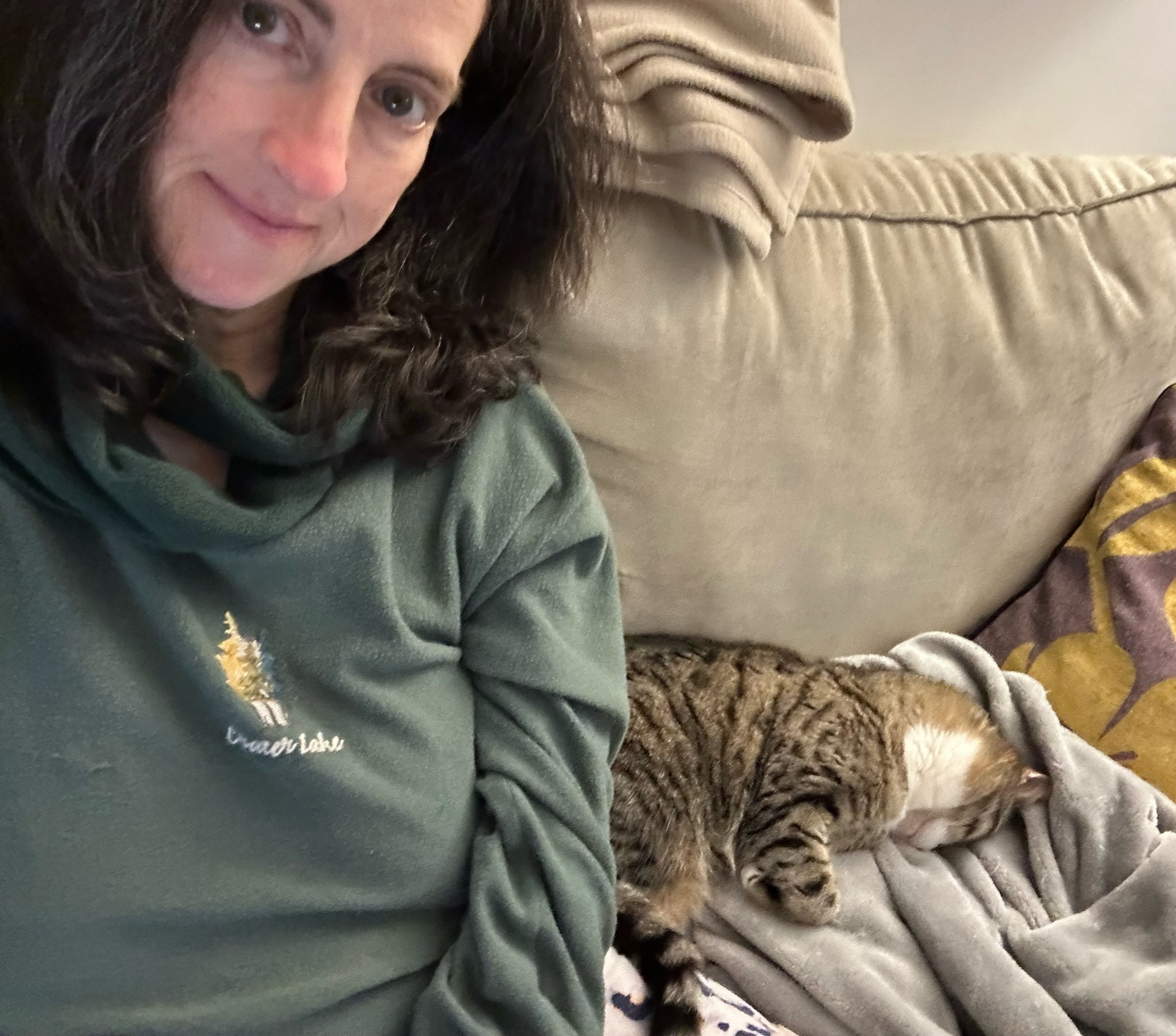 Selfie of myself (white female with brushed out medium length brown hair), wearing a forest green sweatshirt, sitting on a beige sofa. On my left is my cat, gray and white female tabby, somewhat face planted and deeply asleep on a soft gray blanket with a purply and white throw pillow on the other side. My gaze is towards her by way of looking at the camera screen.