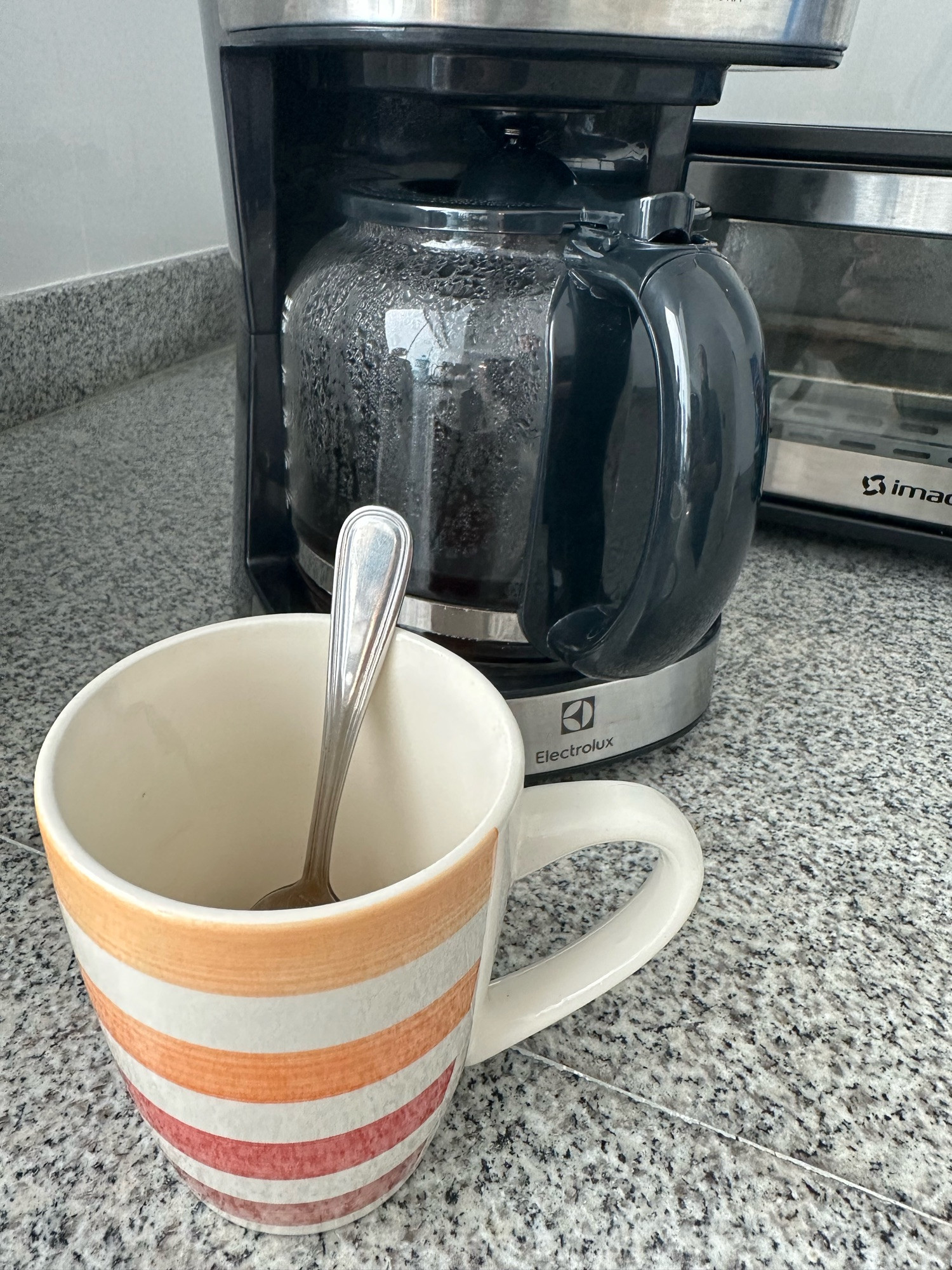 On a black, gray and white speckled kitchen countertop, there is a black and silver 12-cup drip coffee maker having just percolated a couple cups. In front of that is an orange and red horizontally-striped ceramic mug with a teaspoon in it.