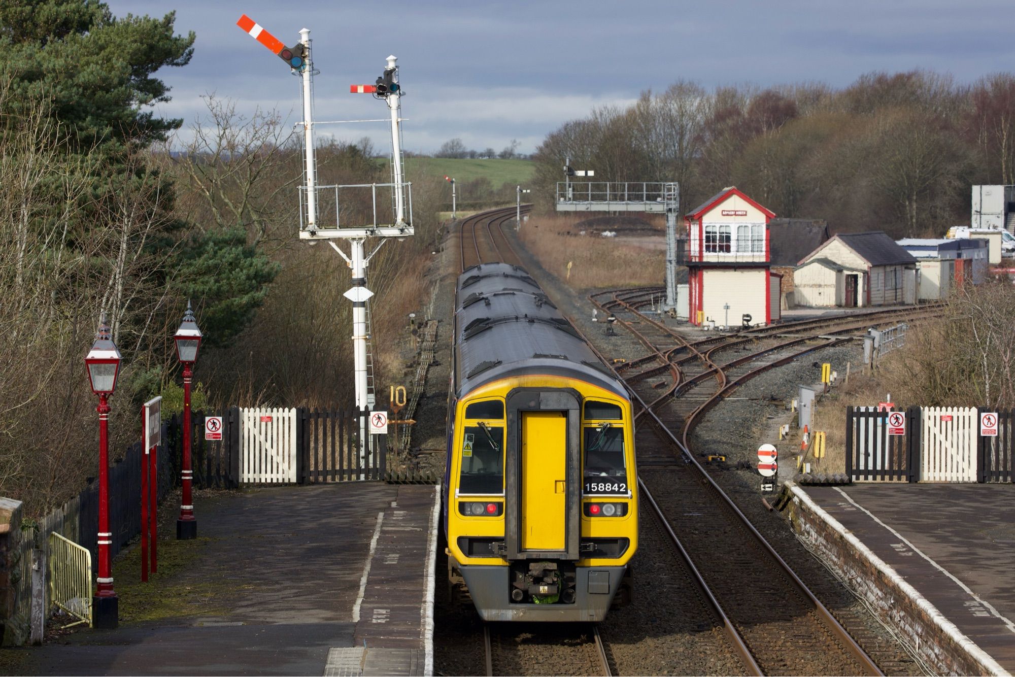 Class 158 - numbered 158842 - departs Appleby station on a Carlisle-bound service passing the tall semaphore signal cleared for its departure. Appleby North signal box is on the right.