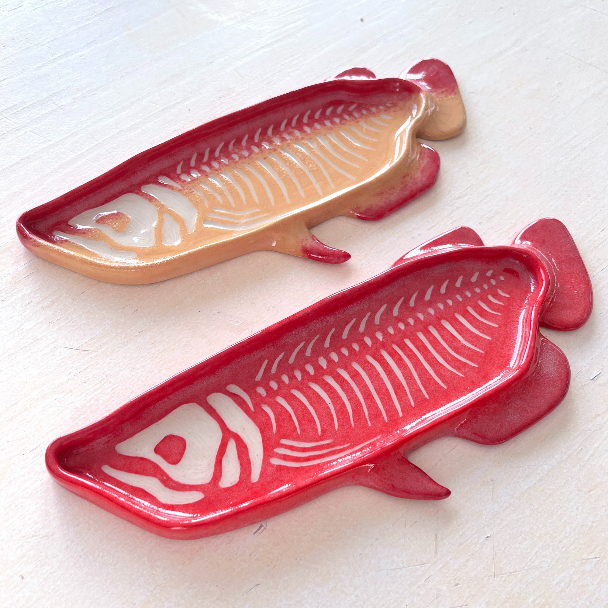 photo of two ceramic arowana plates with sgraffito skeletons carved into the surface. the top plate is red with a yellow belly, and the bottom is entirely red