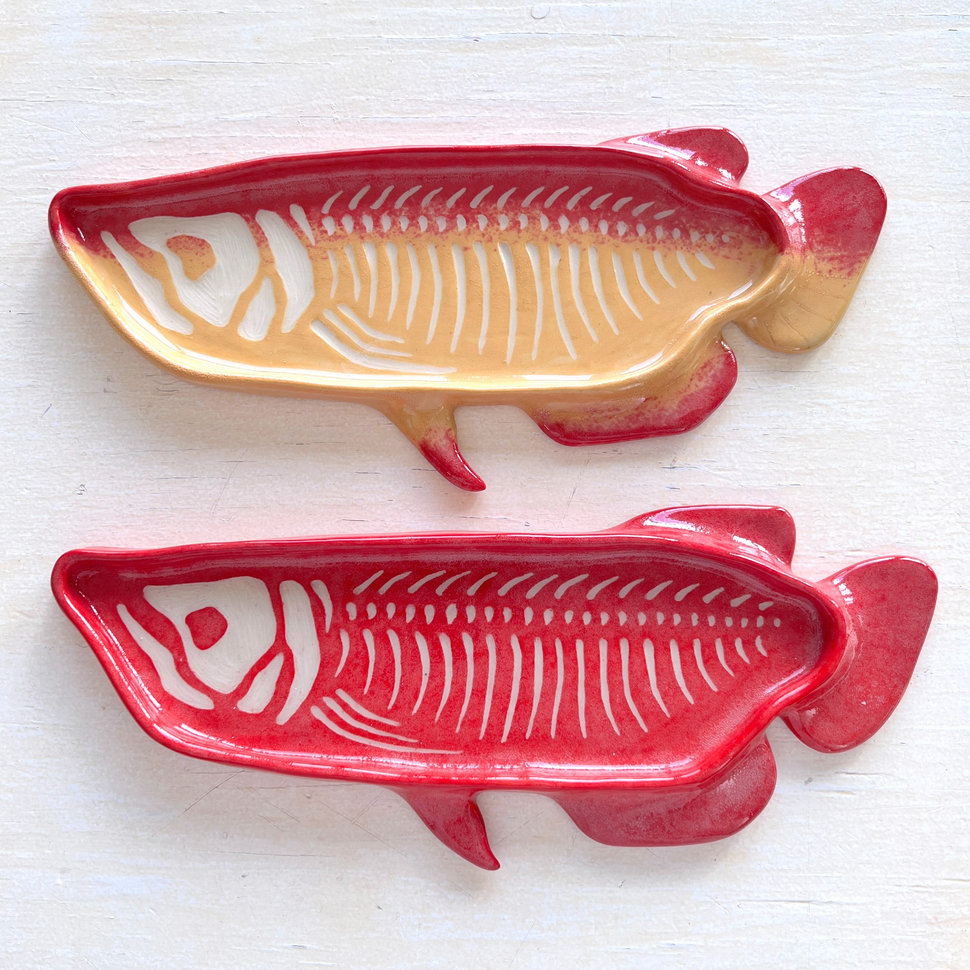 photo of two ceramic arowana plates with sgraffito skeletons carved into the surface. the top plate is red with a yellow belly, and the bottom is entirely red