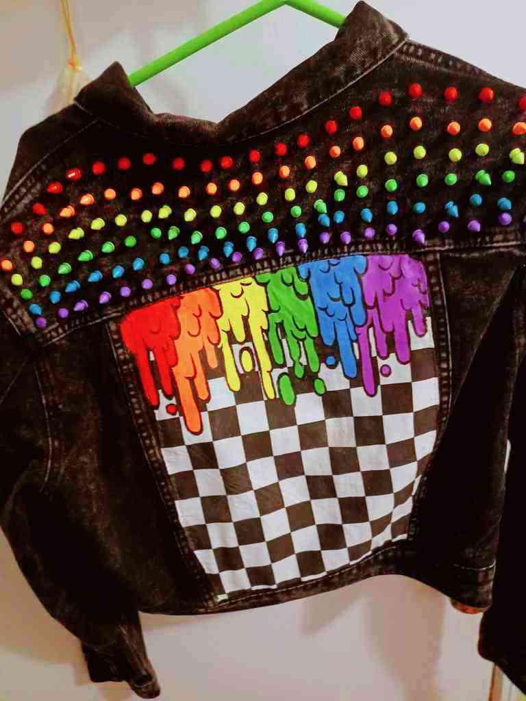 the back of a black denim jacket, there are rows of short tree spikes on the shoulder panel arranged in the 6-color pattern of the rainbow pride flag . the center back panel of the jacket has a panel of checkprimt fabric with goopy design in rainbow colors hand painted onto it.
