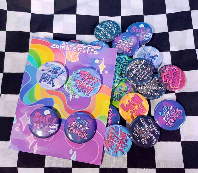 A button set sitting on top of a pile of several more buttons. The backing card has a colorful purple design with a rainbow running through it, there are 4 buttons on it. One says "she/her" with a dark purple background including a ghost and a full moon, one says "zie/hir" with a soft, cloudy background, one says "they/them" with a background that looks like the surface of water, the last one says "punks respect pronouns" on a dark purple background with a pink transgender symbol. These are all in the pile as well, in addition to other pronoun buttons, one that says "two spirit" with a drippy pattern background, and some that say "I'm like stone cold steve austin, I put homophobes in coffins!" on a black background with small rainbow tinted coffin icons.
