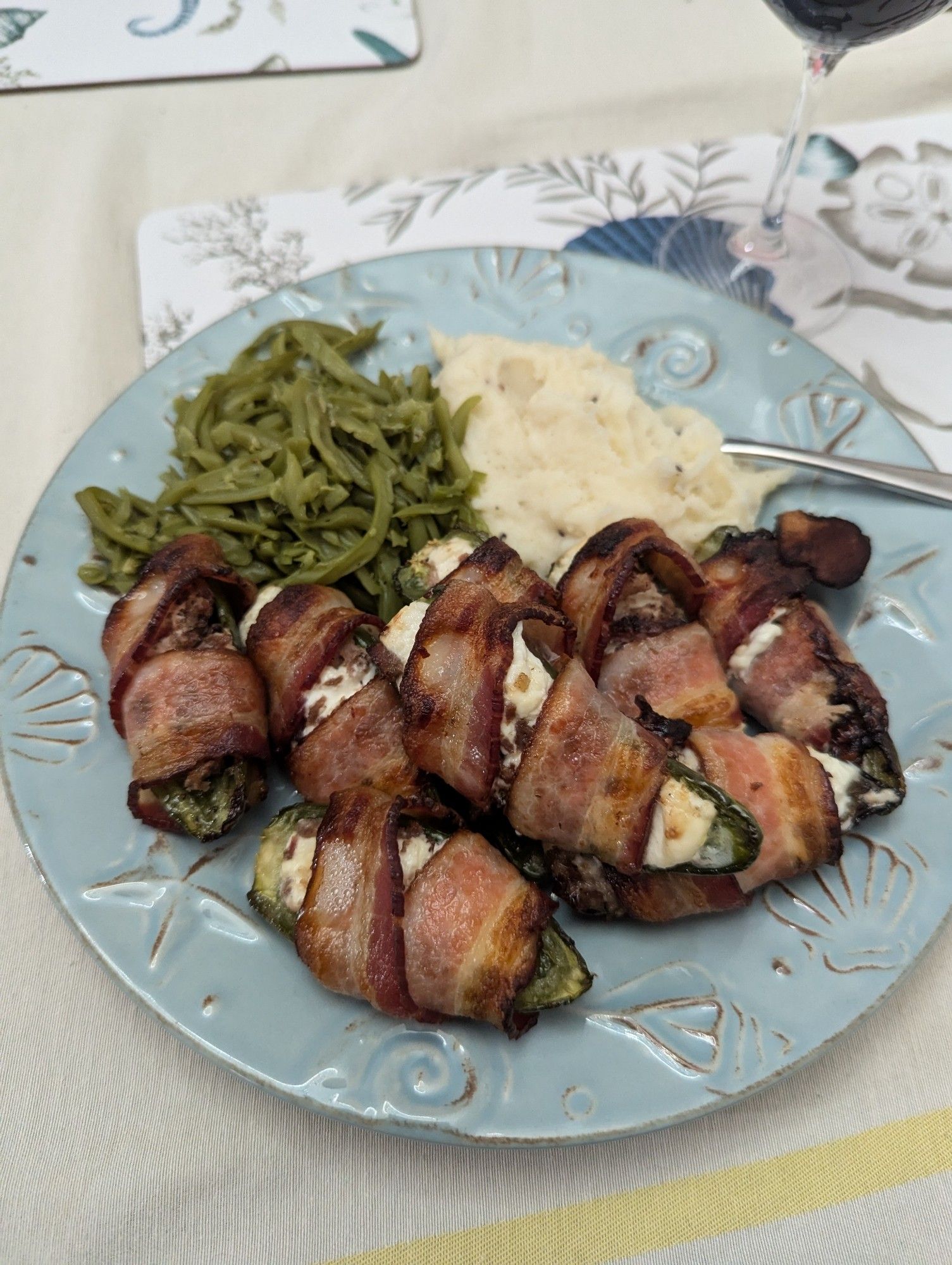 Brisket jalapeno poppers, mashed potatoes, and green beans on a blue plate that has seashell designs around the edge. On top of a placemat that also has seashell designs in blue and grey on a white background that's on a beige table cover with stripes around the edge.