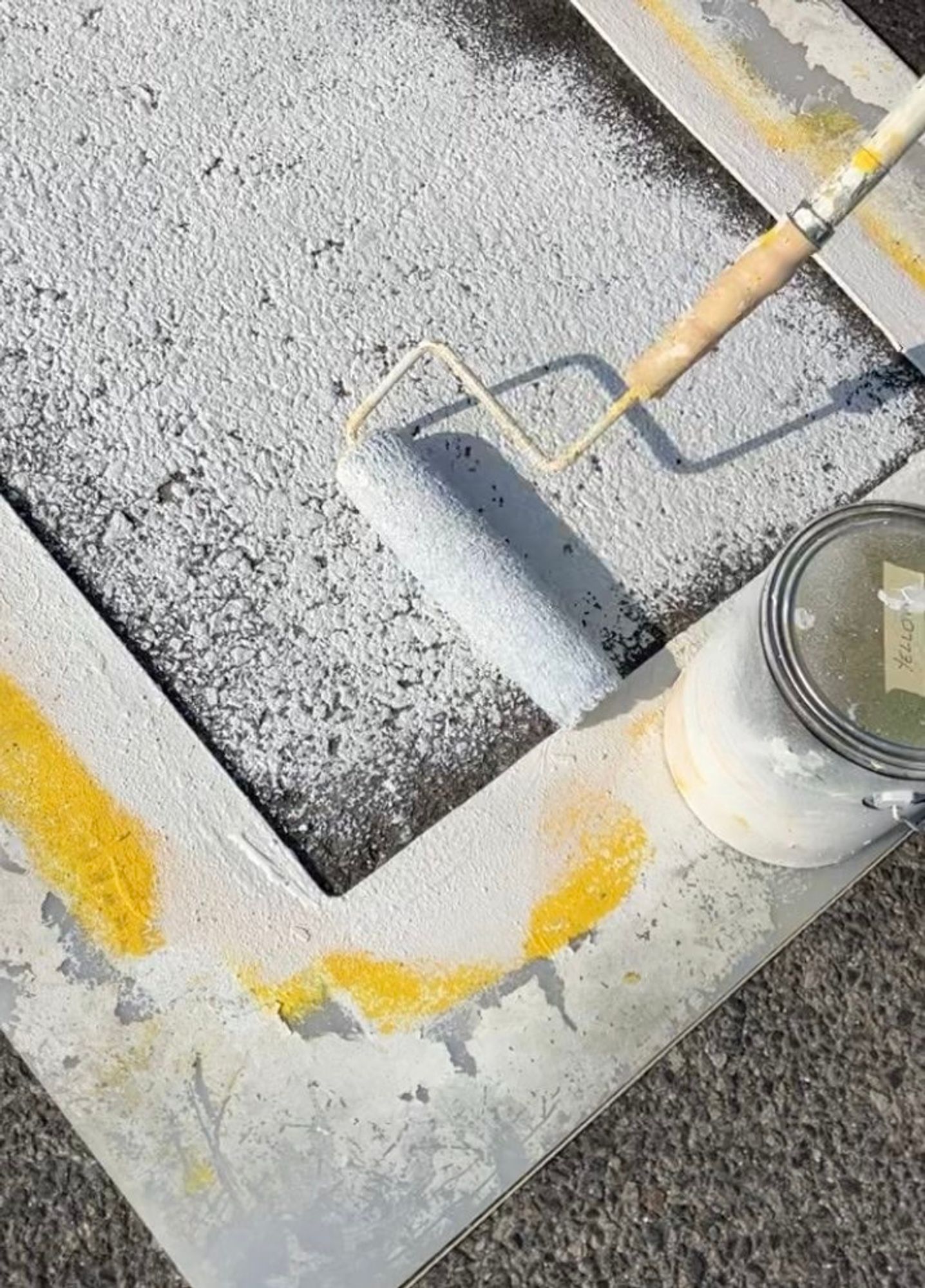 A crosswalk stencil held down by a can of paint with a paint roller in the center rolling out white paint. 