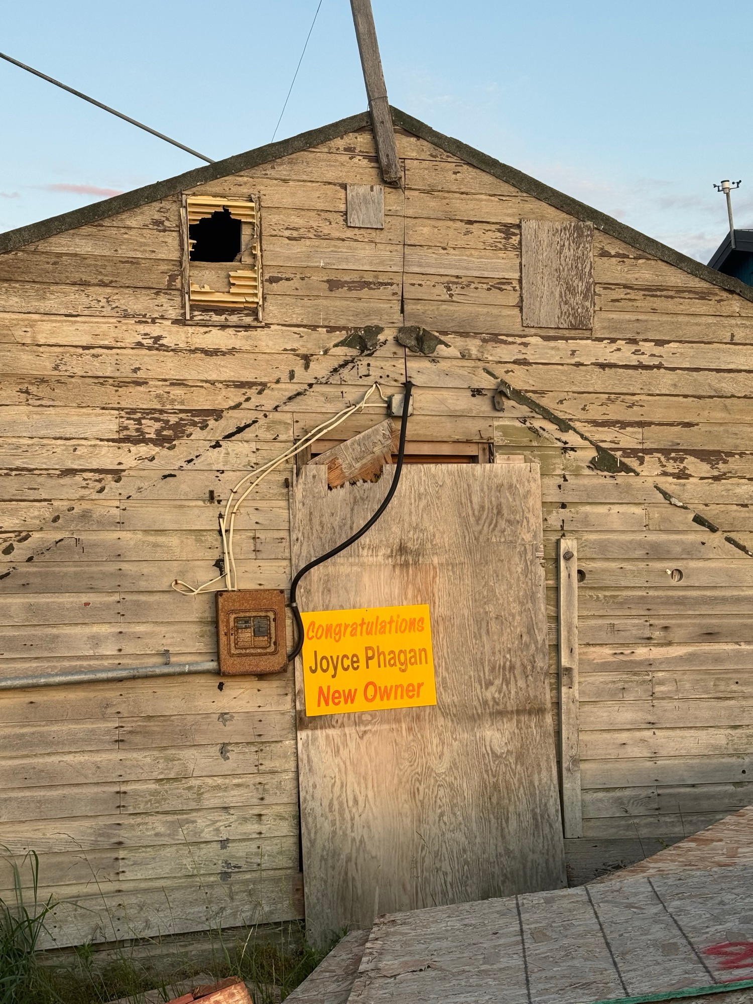 An old weathered building with no paint and a broken vent. A bright yellow sign on the locked door reads “Congratulations Joyce Phagan New Owner”