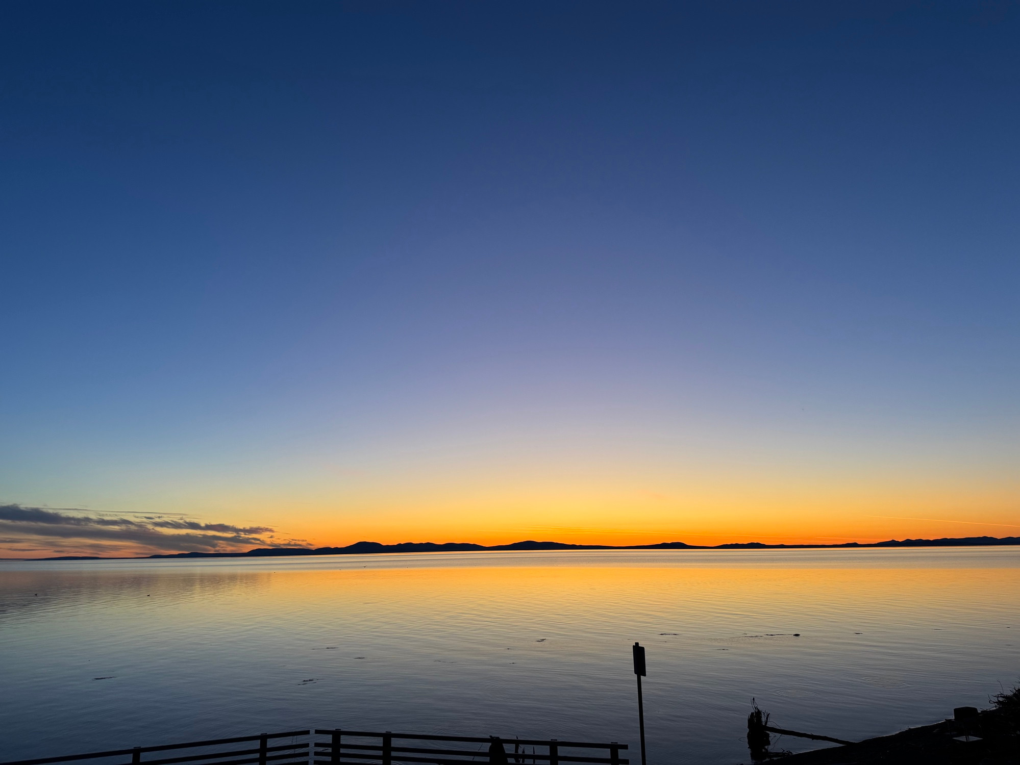 A twilight photo of Kotzebue Sound. The  water is still as glass and the distant hills across the sound are framed in yellow orange glow accentuated by smoke from tundra fires in northern Alaska. The sky is almost gloaming blue.