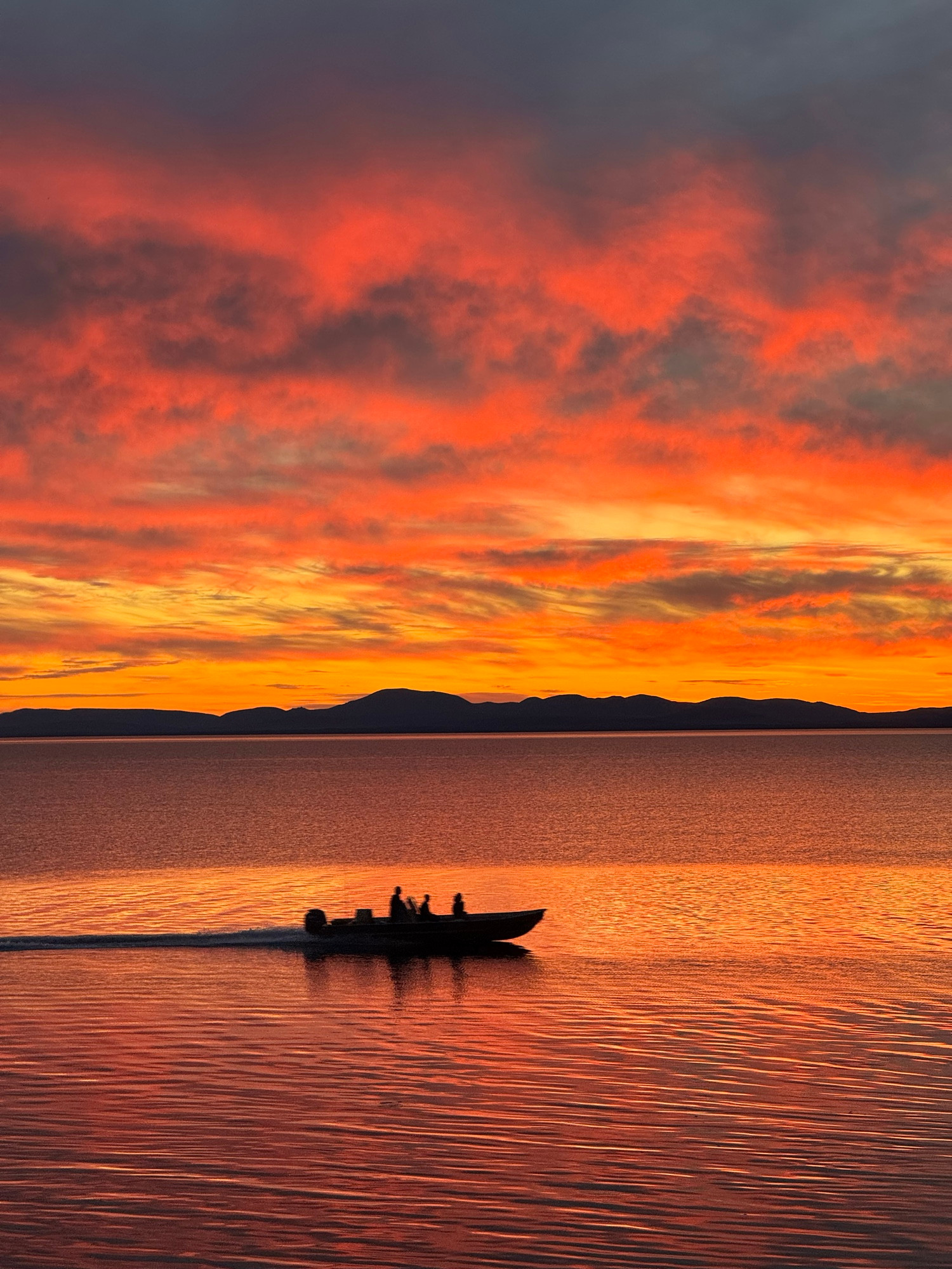 A silhouetted fishing boat crosses glassy clear Kotzebue Sound at 1:00am twilight. The clouds in the sky are ablaze in the glow of the recently set sun.
