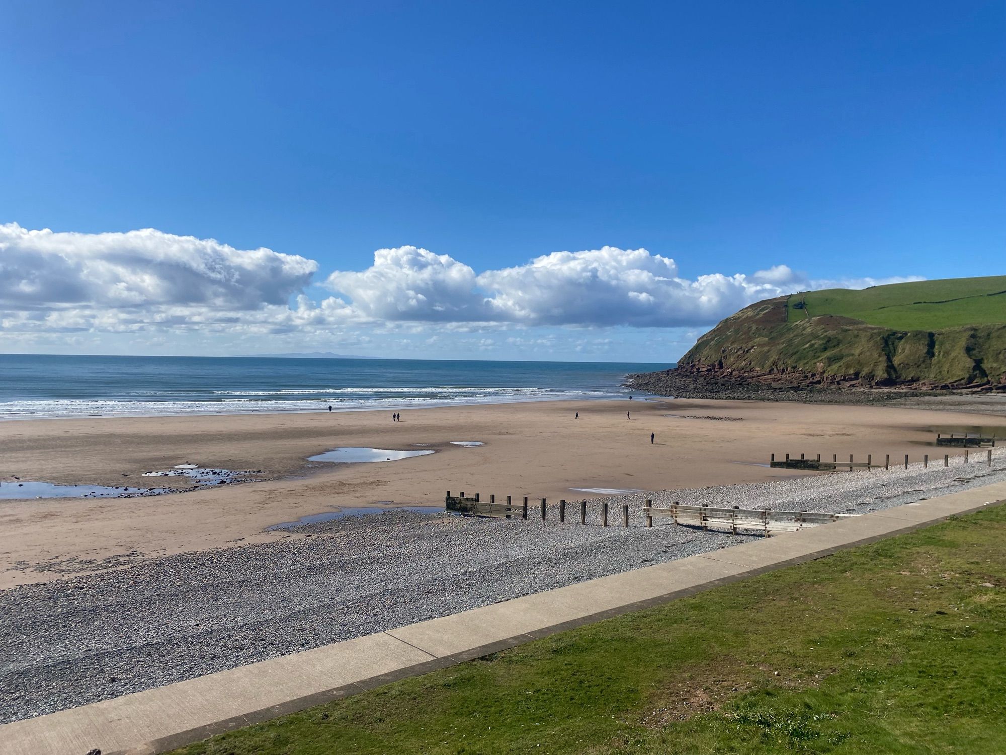 A view across a sandy beach towards a calm sea below a very blue sky. Clouds are just about the horizon and there are low cliffs to the right. There is pebbly shingle at the top of the beach