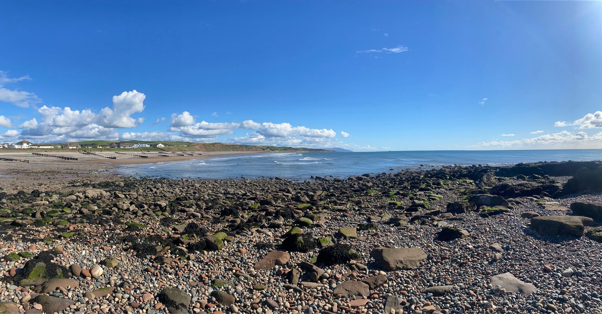 A view across a rocky foreshore towards the sea beneath a dazzlingly blue sky dotted with light, white clouds