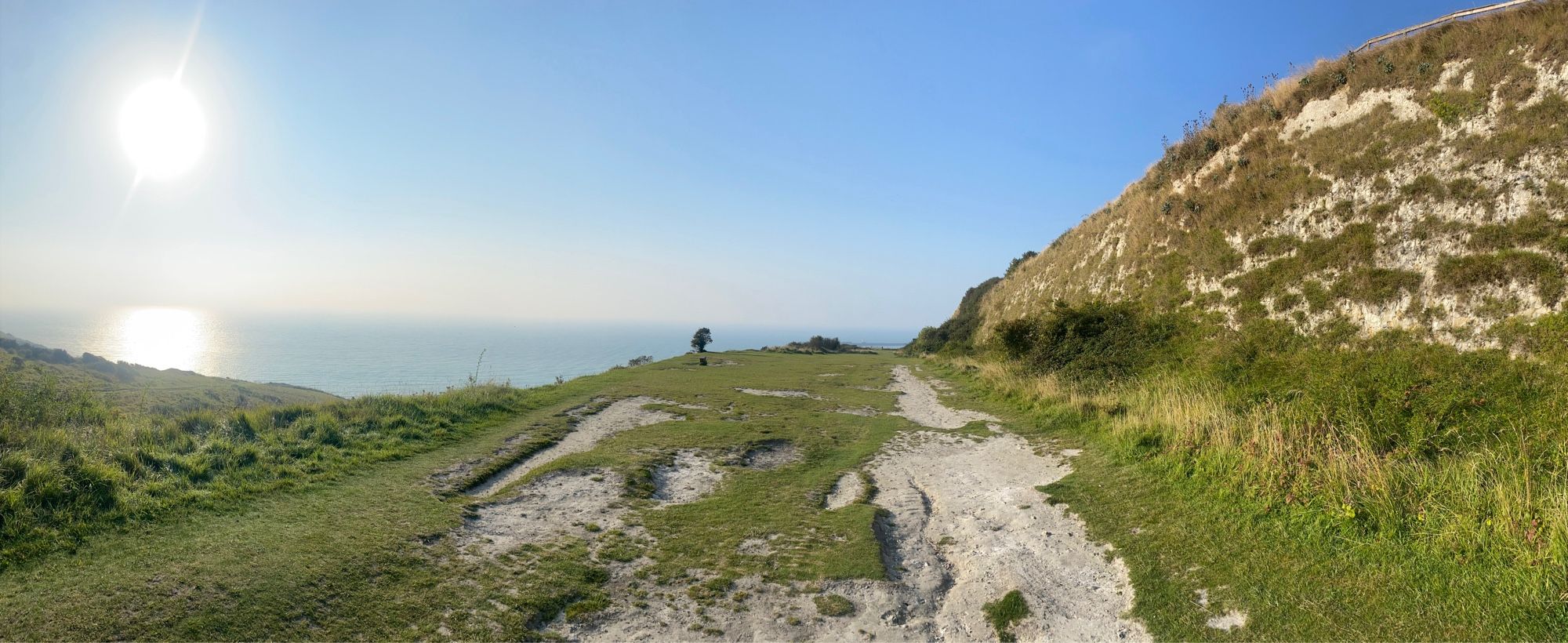 A view out to see below a low, rising sun in a blue, cloudless sky. In the foreground is a white rough track cutting across a grassy ledge. There is a white, slightly grassy slope on the right of the shot