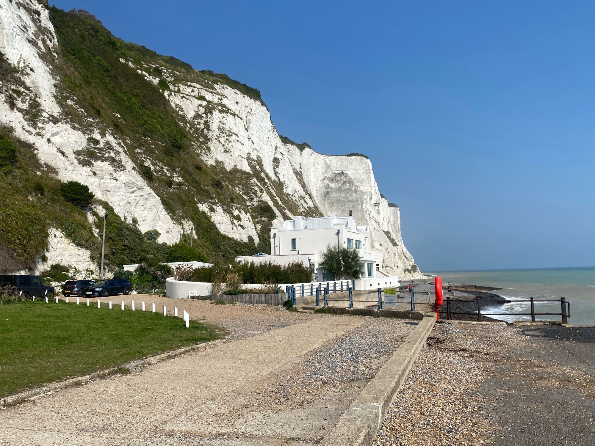 White chalk cliffs, about 100 m tall, next to the sea. The sky is blue and cloudless. In the middle ground is a white, art deco house. In the foreground is a pebble beach and a beach side path.